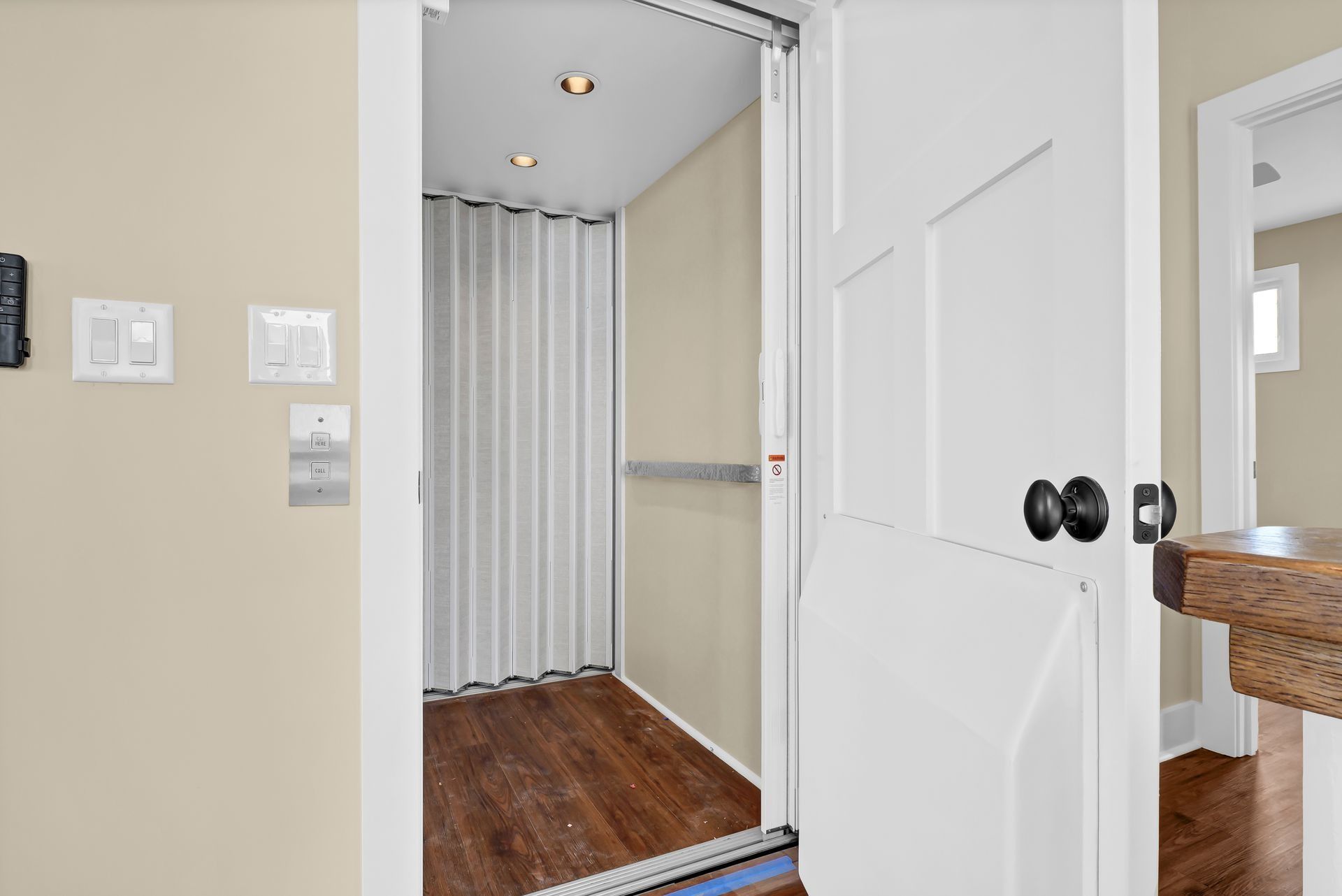 Elevator doorway with white accordion door and wood floors. Beige walls. Light switches visible.