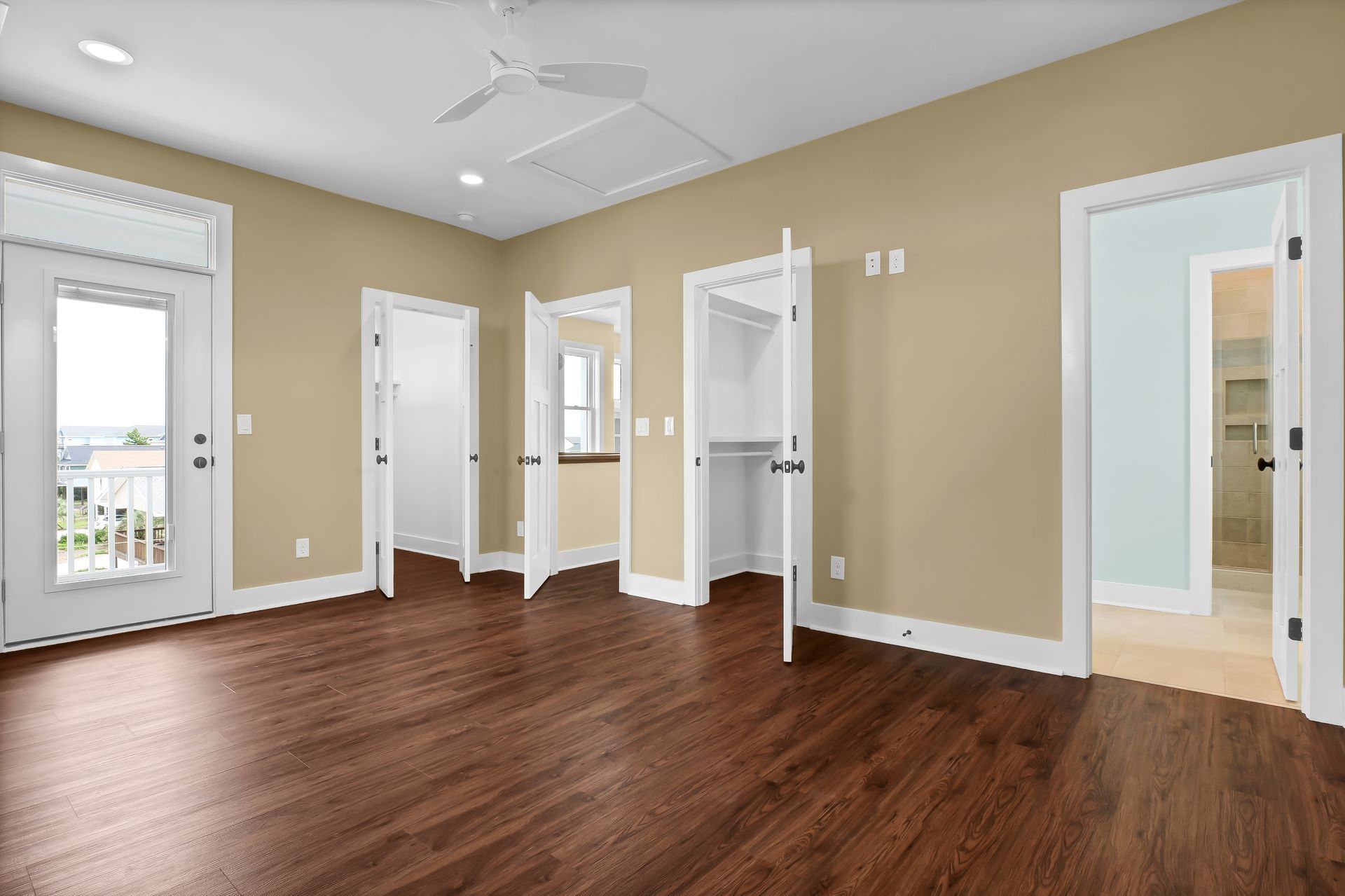 Empty bedroom with wood floor, beige walls, and white trim. Doors to balcony, bathroom, and closets.