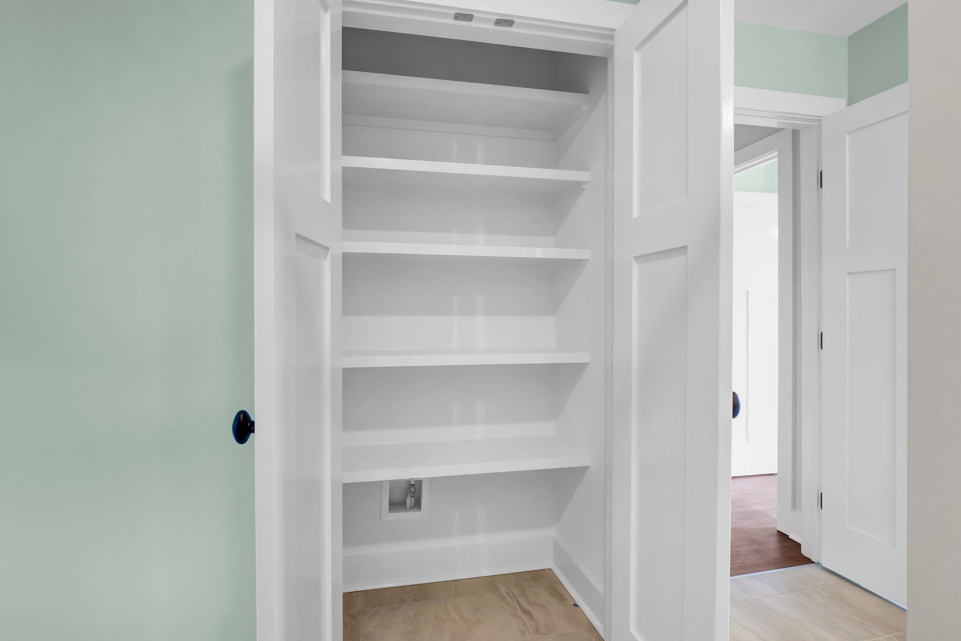 White pantry with open doors, multiple shelves, and a power outlet, situated next to a light green wall.