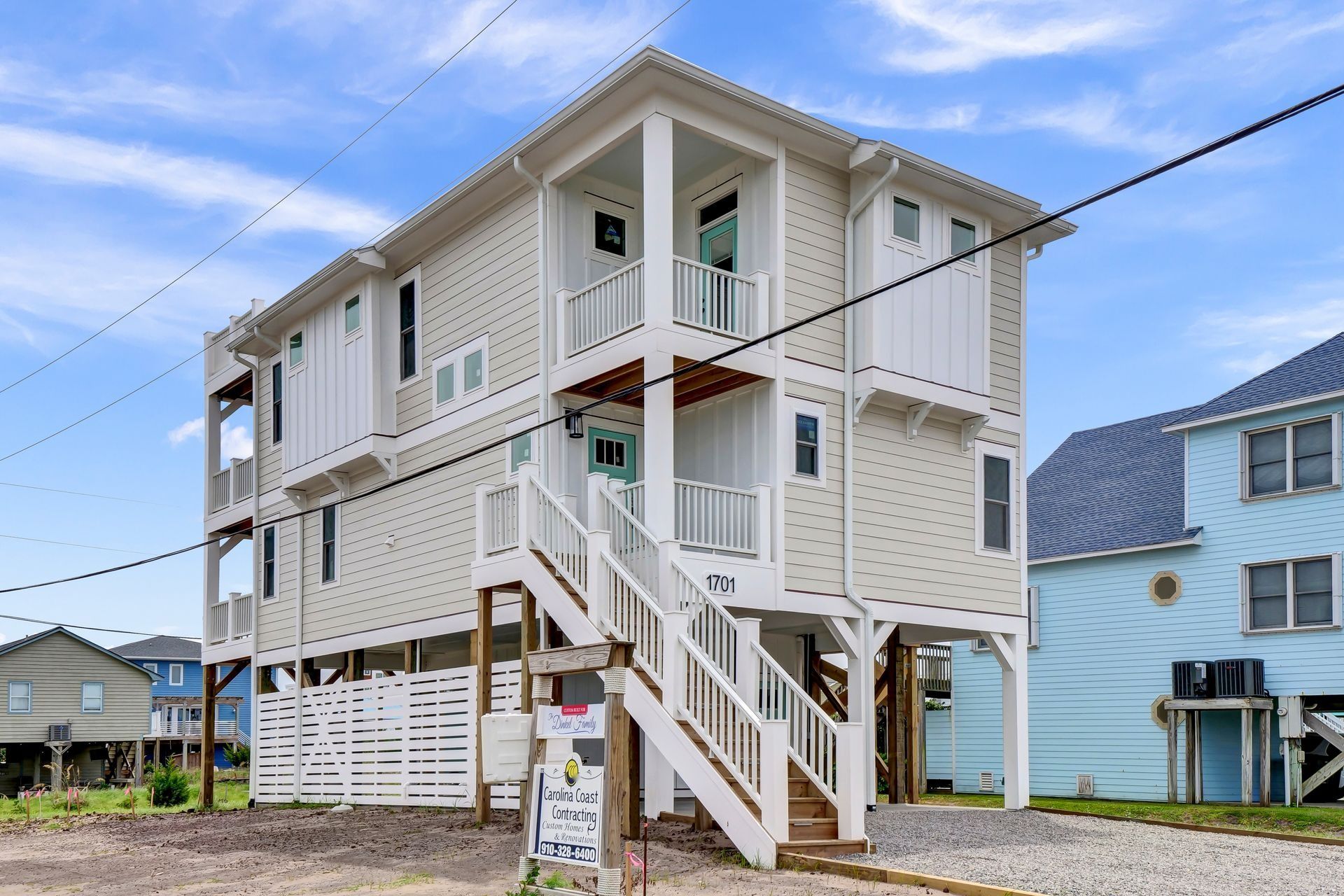Two-story beach house on stilts with white and light tan siding and wooden stairs. Blue sky.