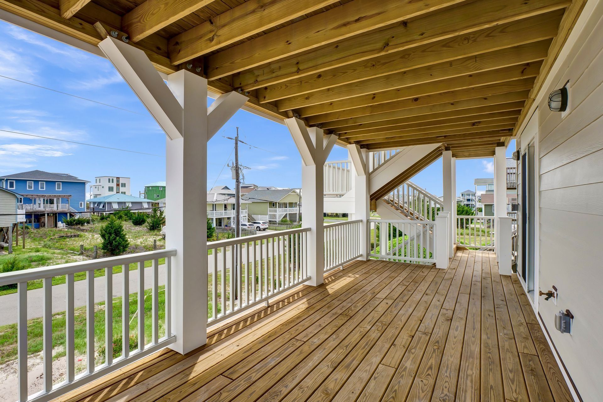 Covered wooden deck with white railing, posts, and stairs, overlooking neighborhood under blue sky.
