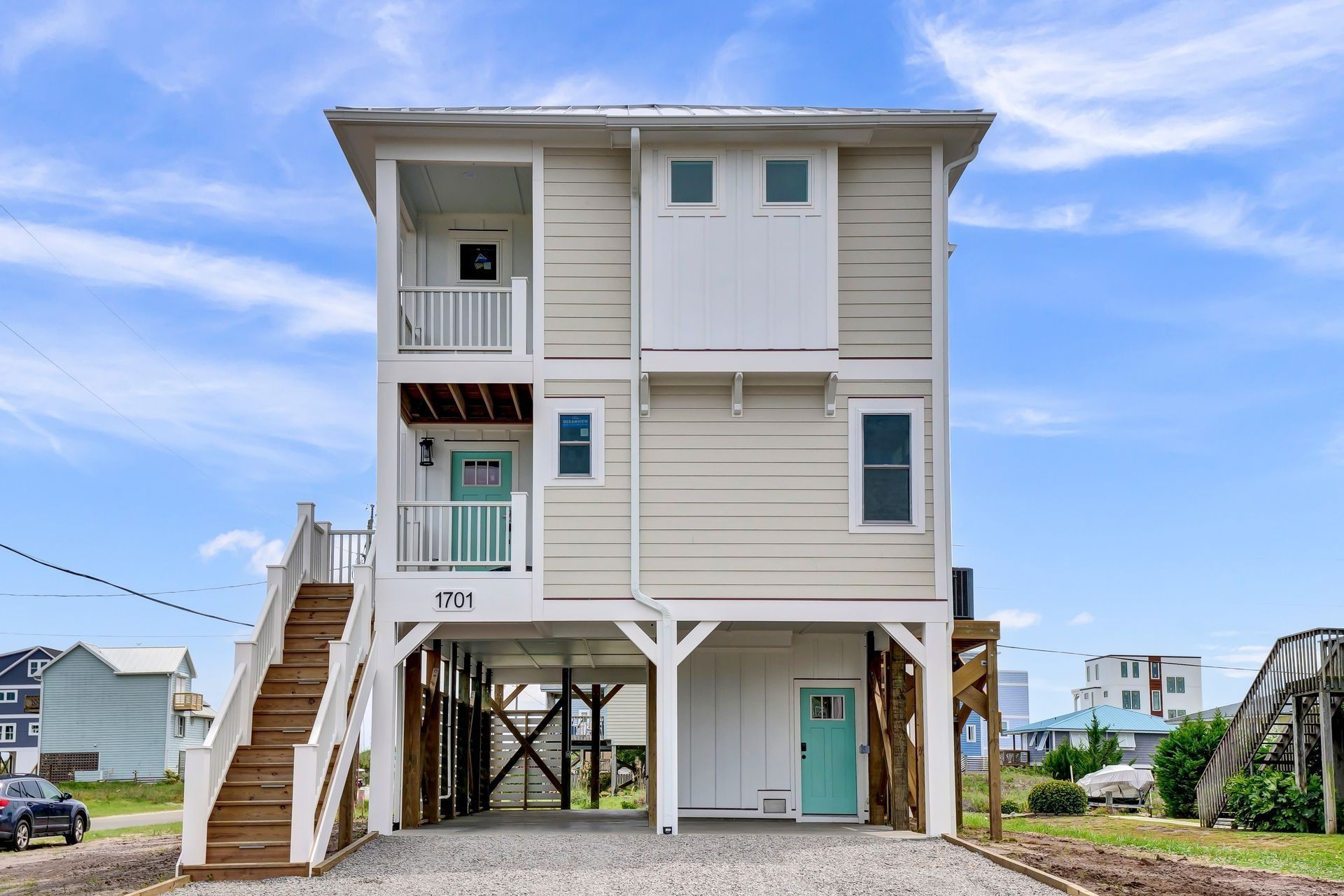 Two-story beach house on stilts with wooden stairs, light siding, and turquoise doors.