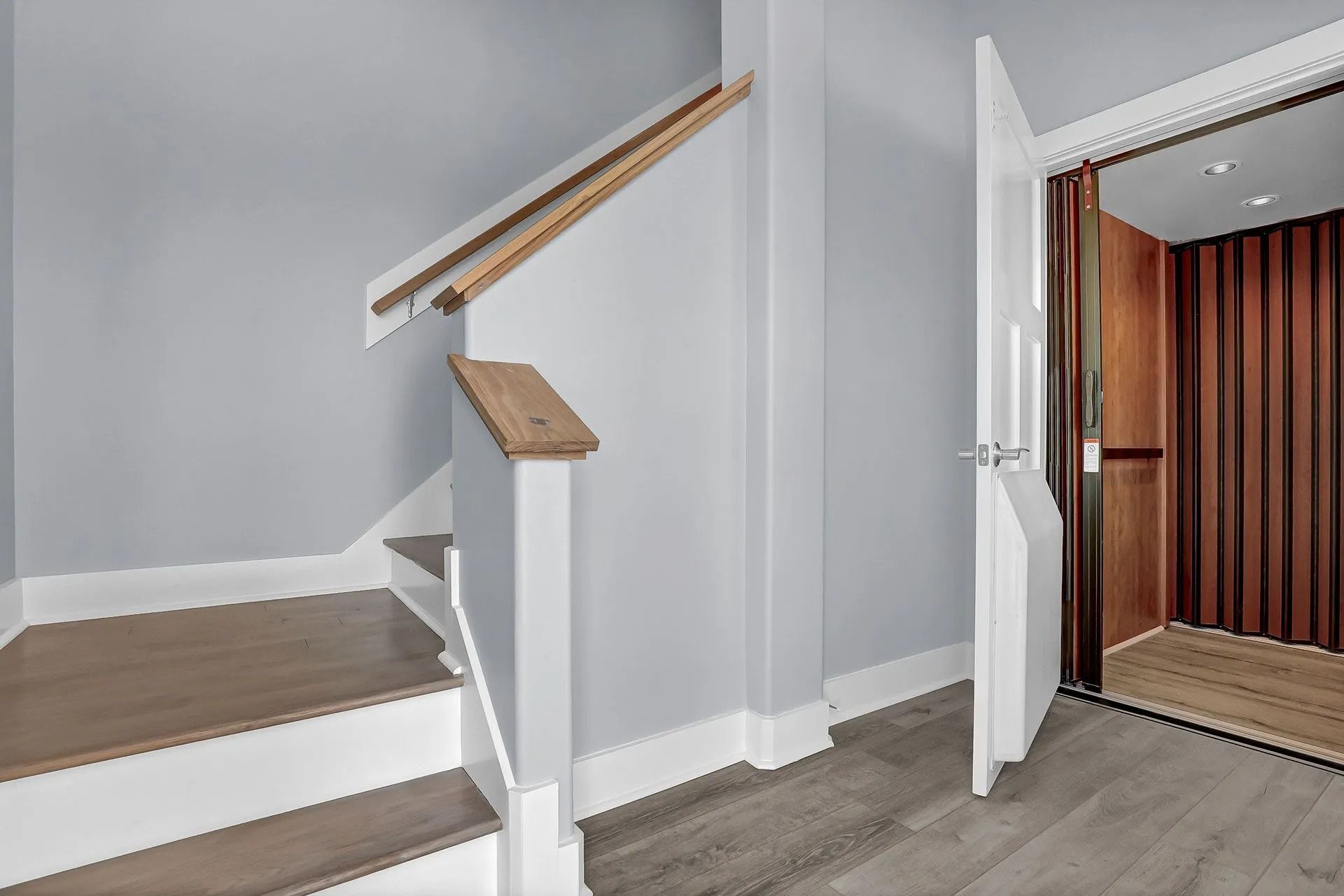 A staircase and a home elevator in a light gray hallway. The elevator door is open.