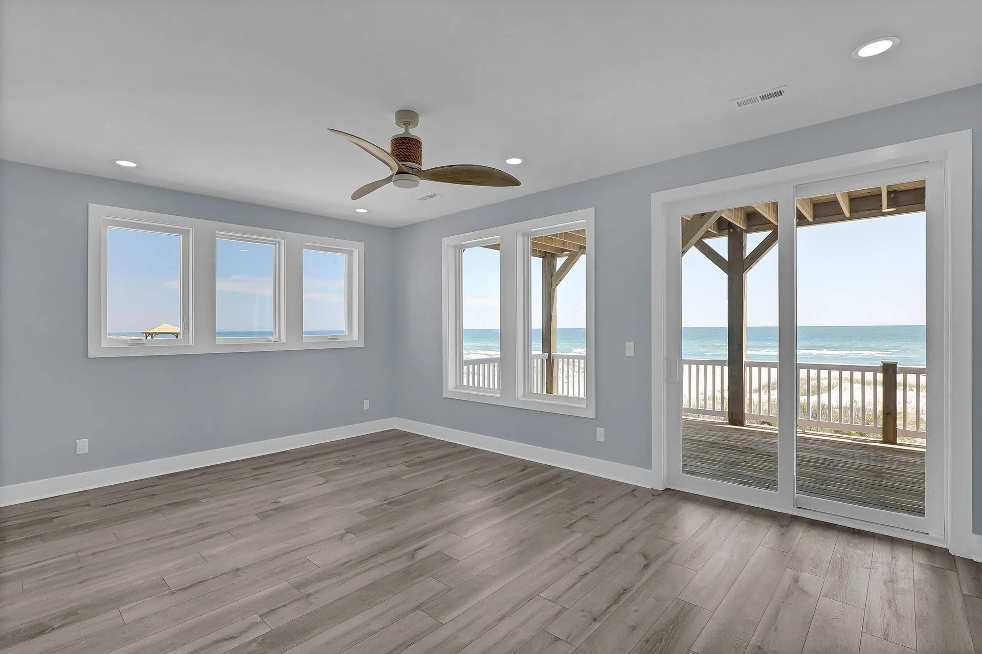 Empty bedroom with ocean view, windows, and sliding doors. Light blue walls, wood floor, and ceiling fan.