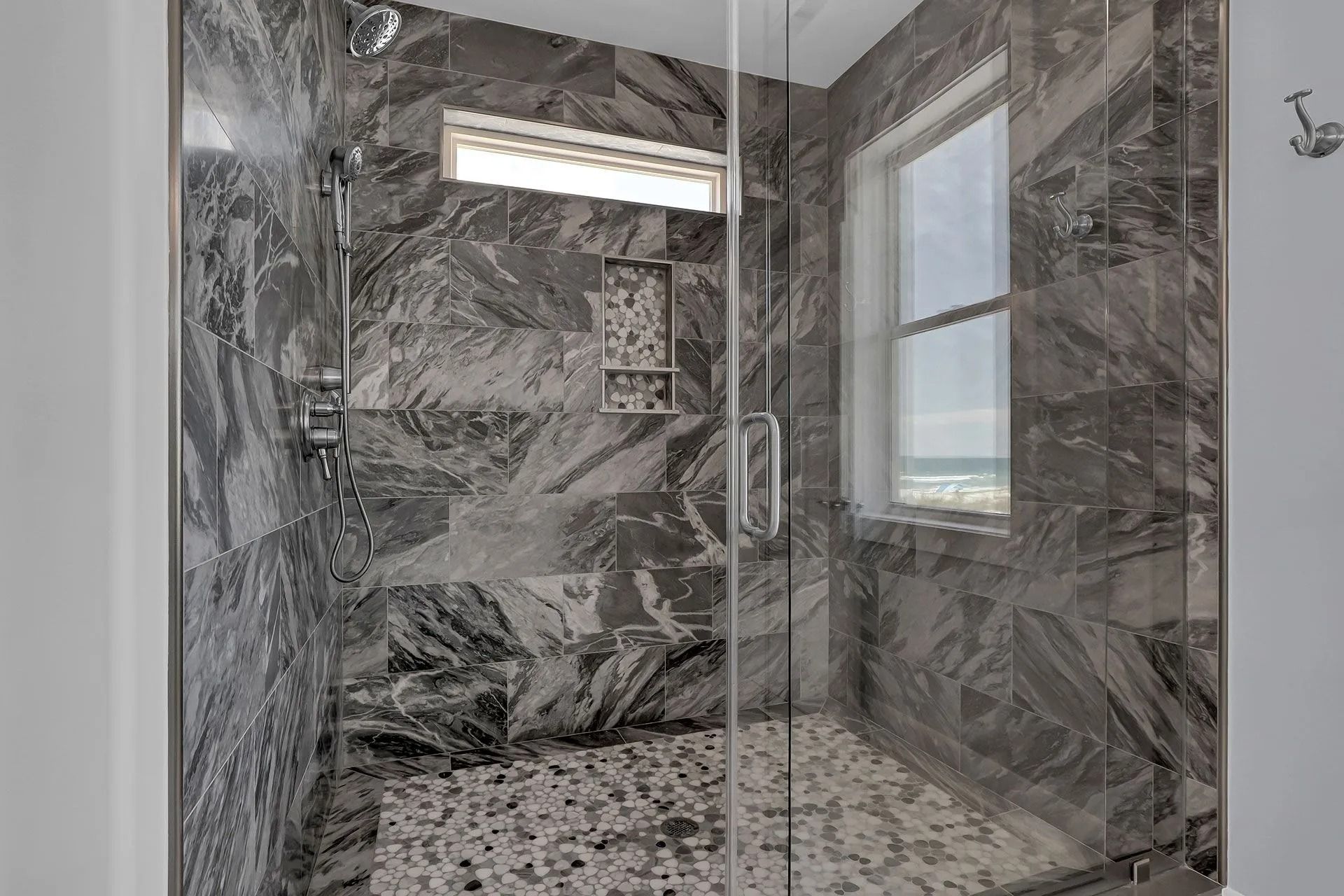 Shower with gray marble tile walls, clear glass door, pebble floor, and a small window.