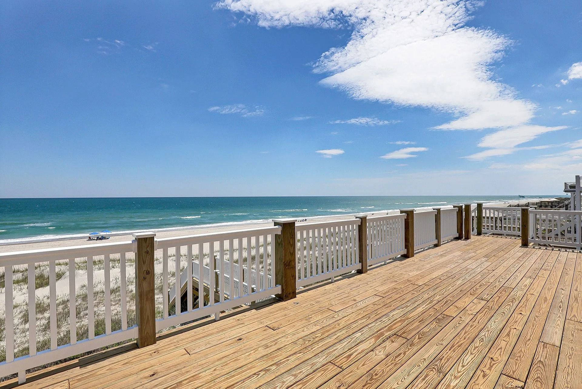 Wooden deck with white railing overlooking a beach and ocean under a blue sky with clouds.