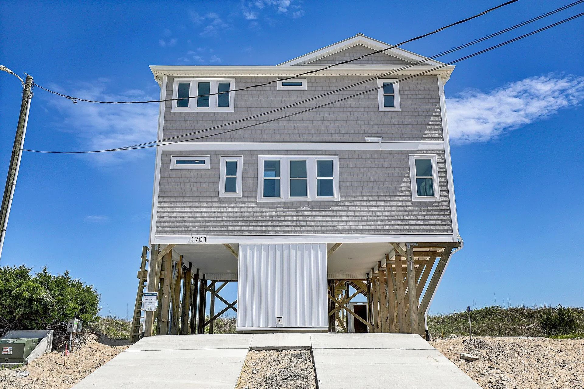 Gray beach house on stilts with a concrete driveway and clear blue sky.