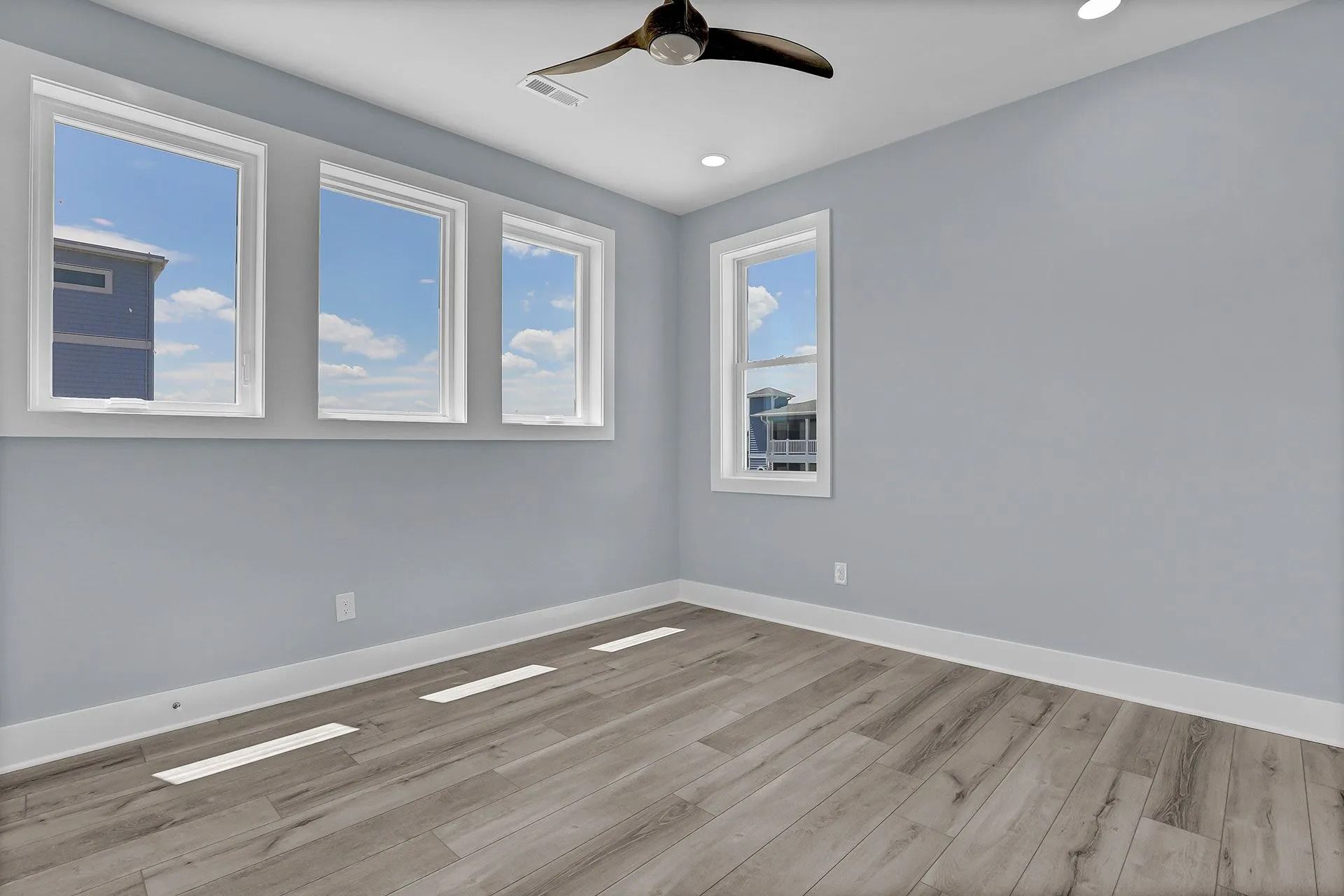 Empty room with blue-gray walls, multiple windows, and wood-look flooring. A ceiling fan is visible.