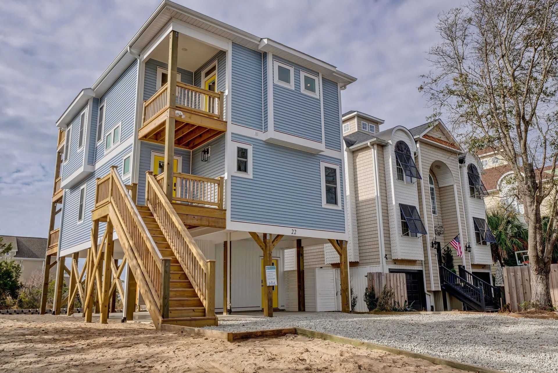 Two-story beach house on stilts, light blue siding, wooden stairs, and a gravel yard.