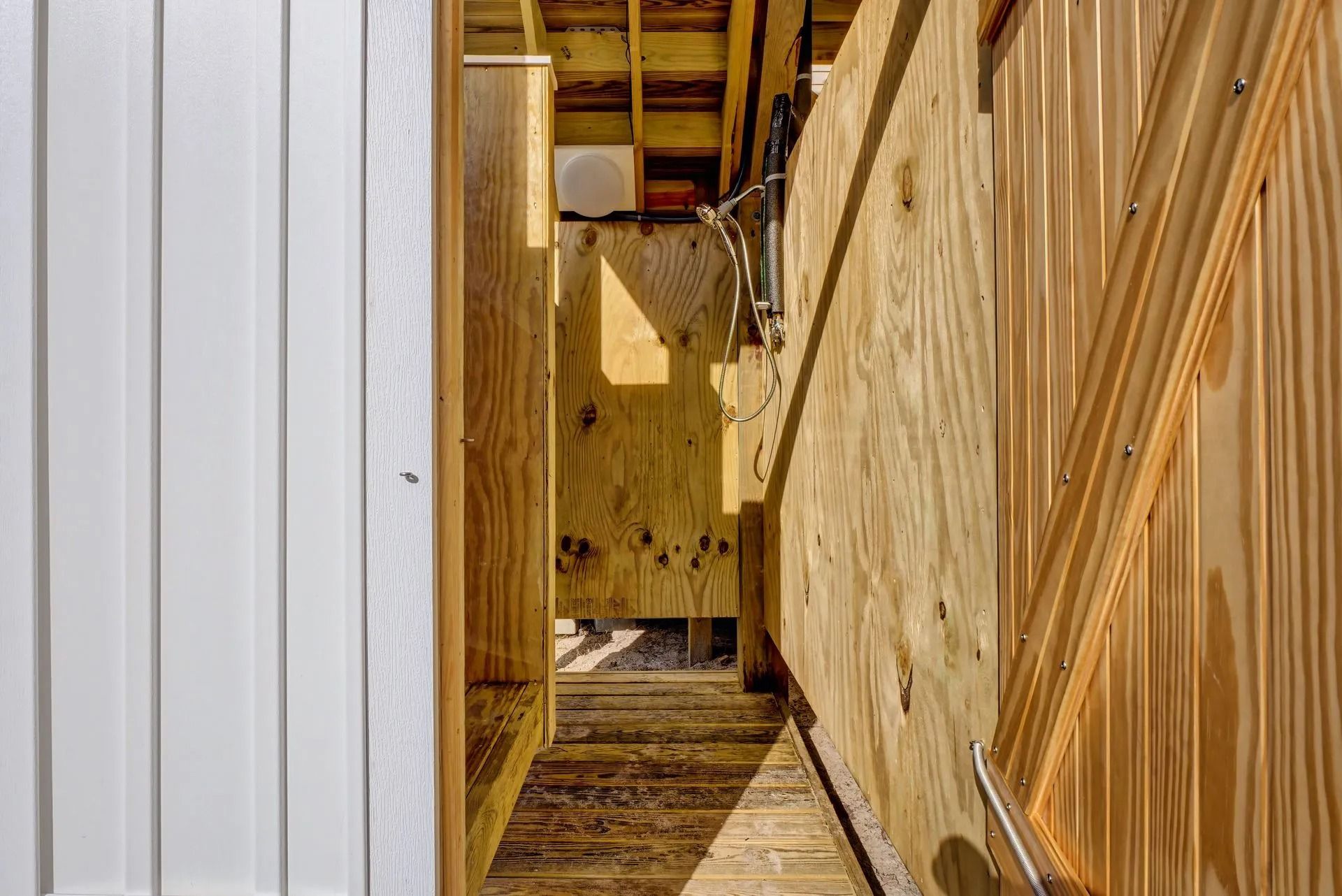 Narrow wooden corridor with plywood walls and partial door. Sunlight shines from the end.