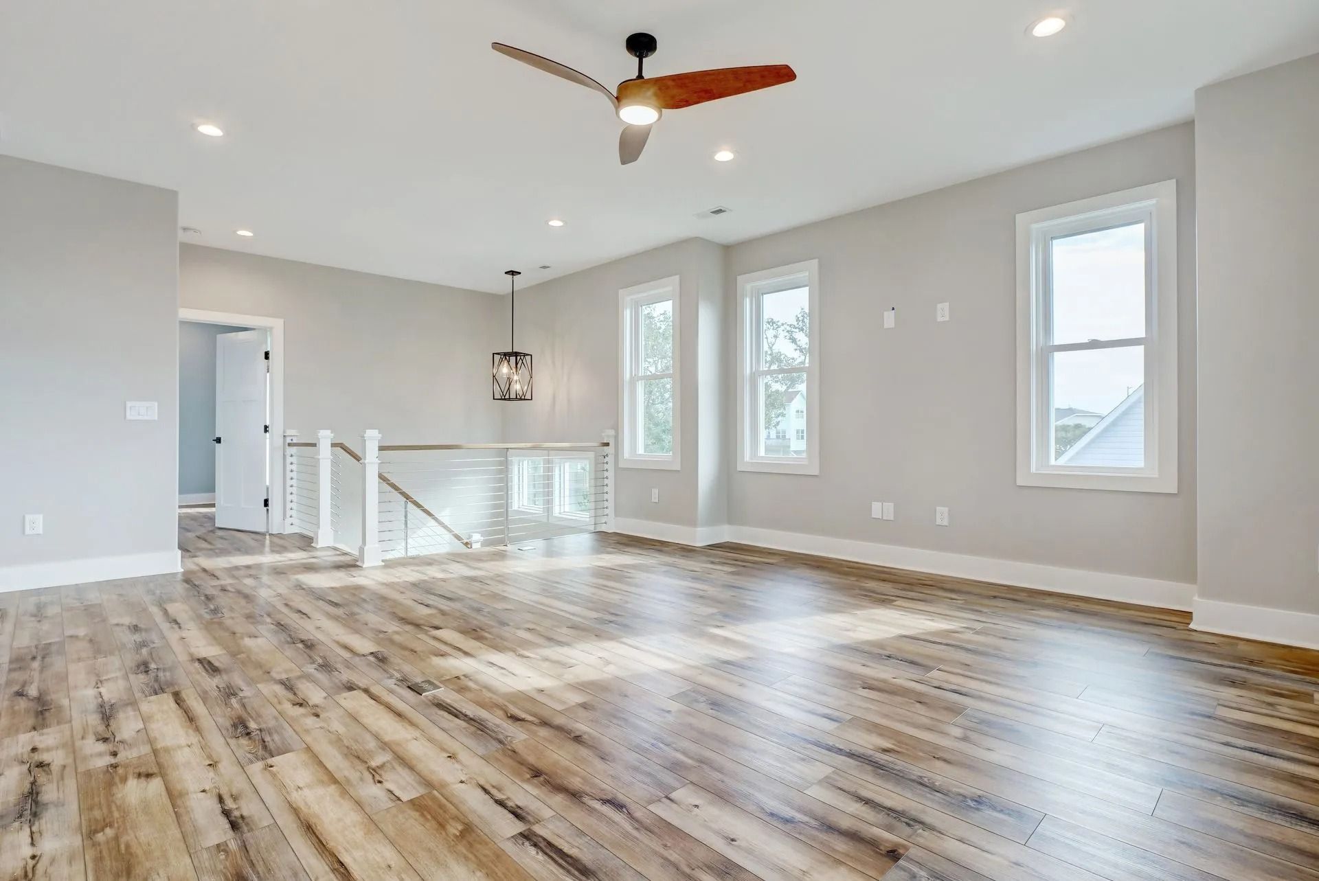 Spacious, light-filled room with wood flooring, three windows, ceiling fan, and staircase leading to a doorway.