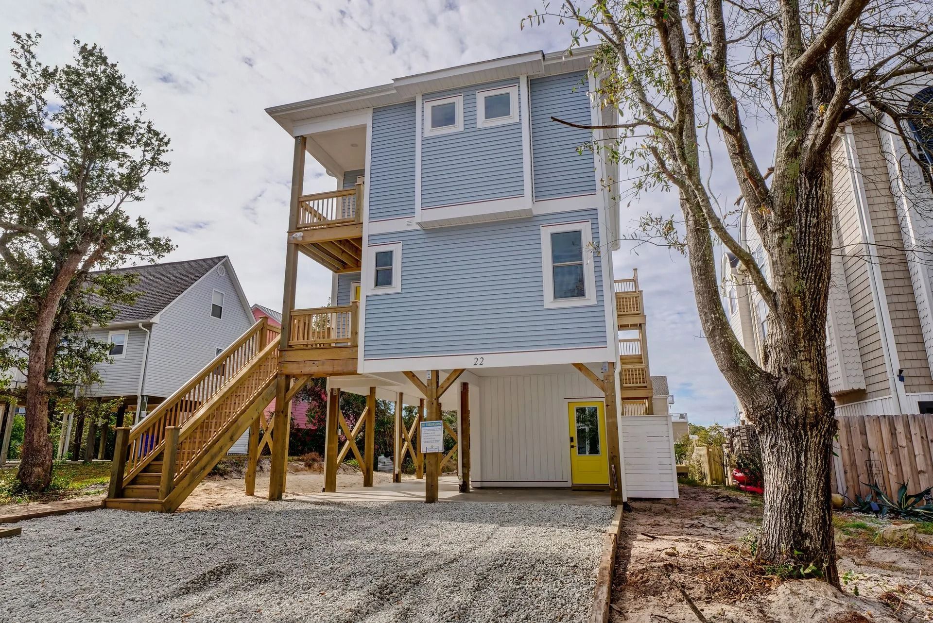 Two-story blue beach house on stilts with wooden decks and yellow door. Gravel driveway, trees.