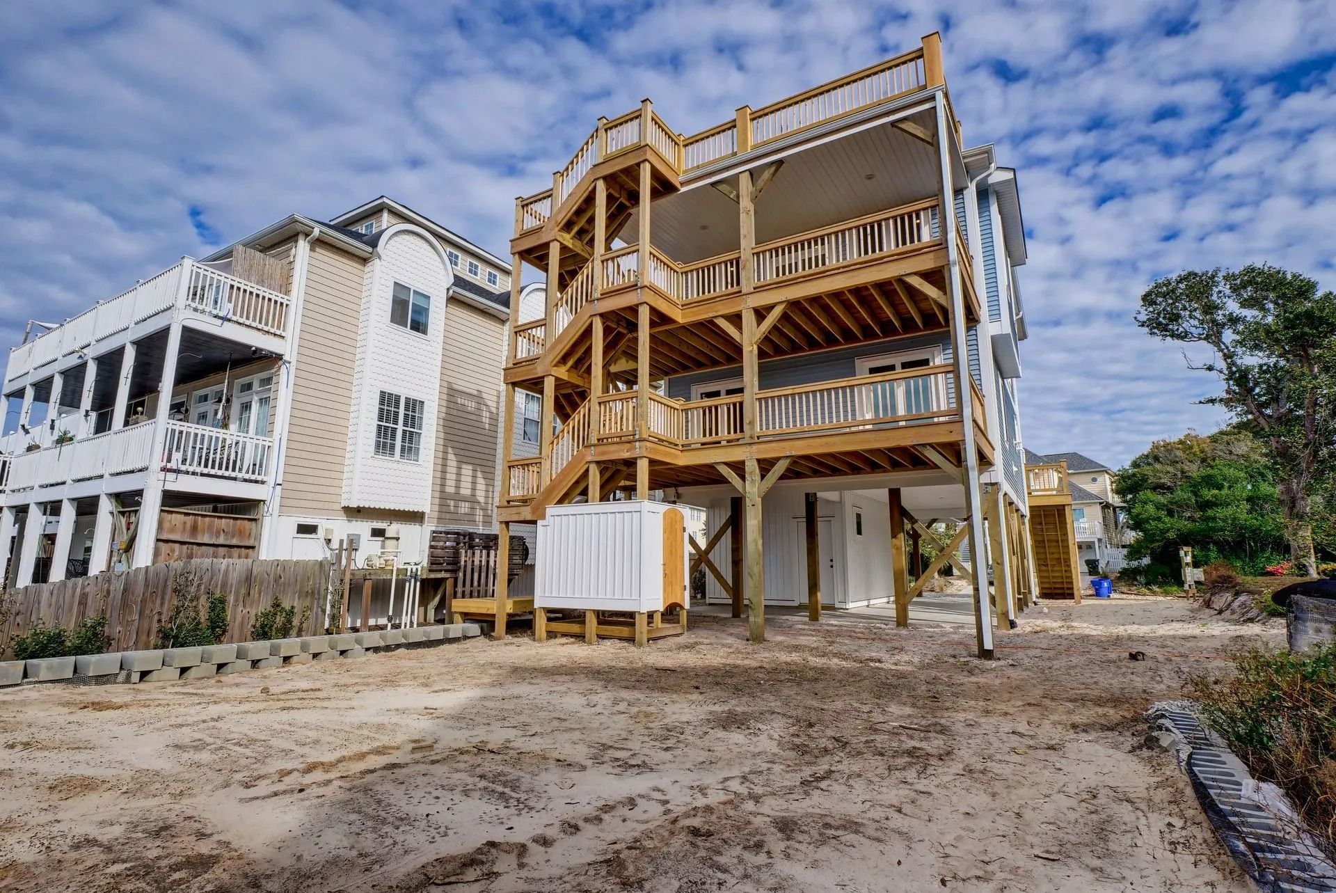 Multi-story wooden beach house with open staircases and deck, on a sandy lot.