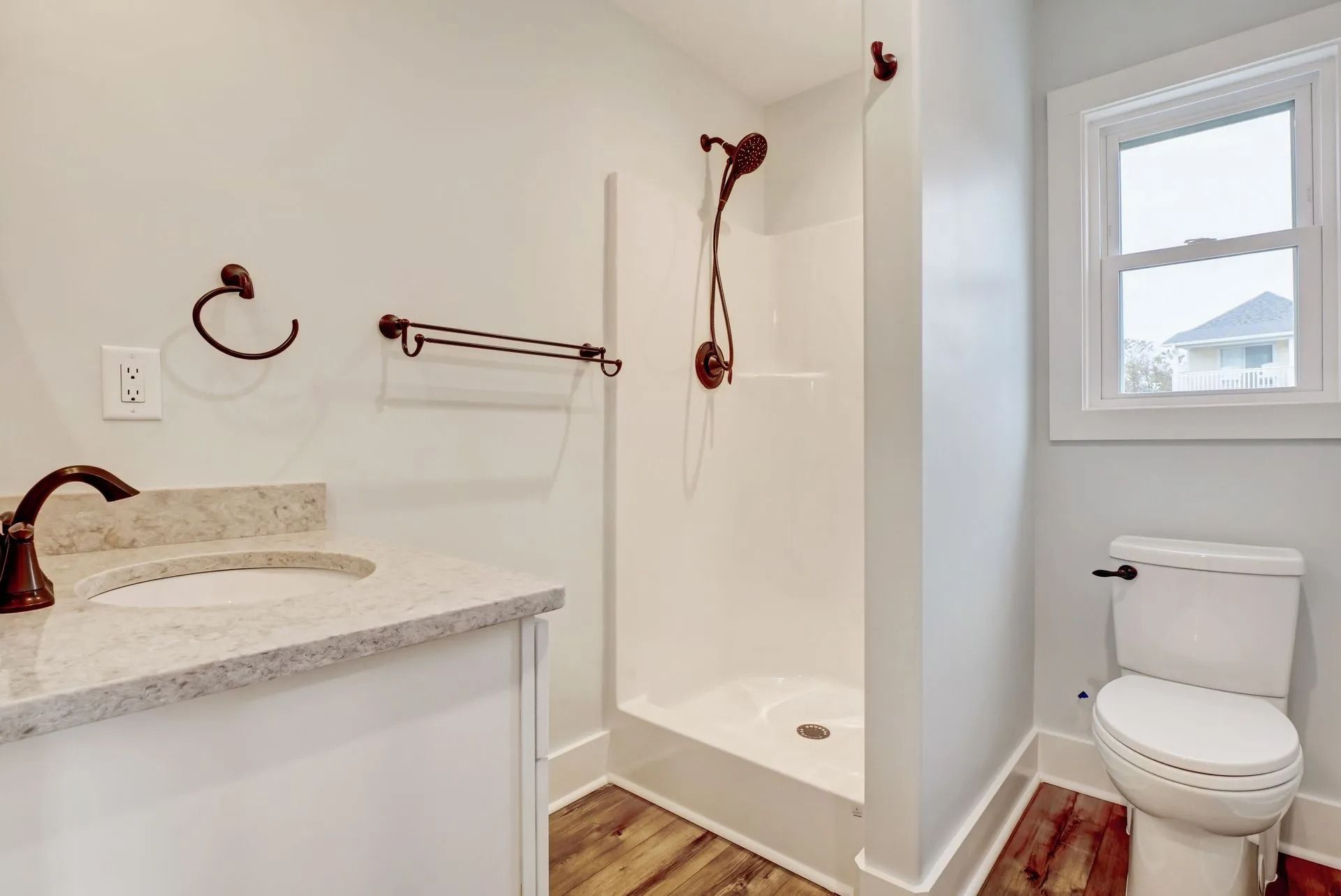 Bathroom with white walls, vanity, shower, and toilet. Brown fixtures and wood-look flooring. Window on the right.