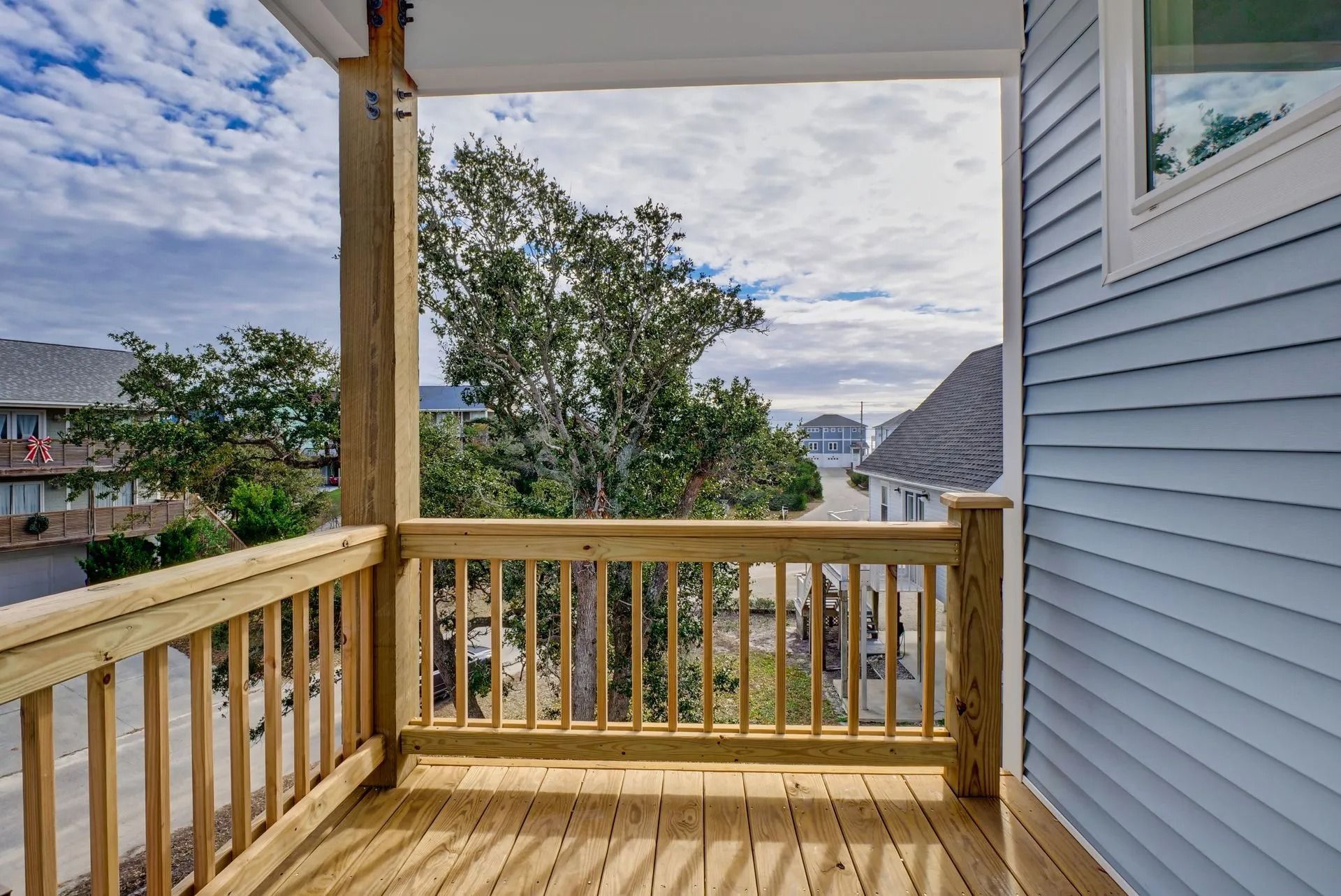 Wooden deck with railing overlooking a street, blue house exterior on the right, trees and sky visible.