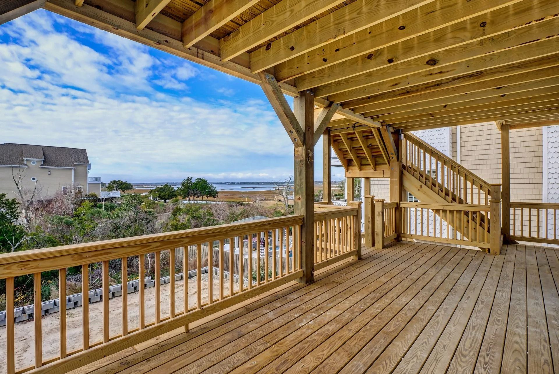 Wooden deck with railing, stairs, and partial view of houses, trees, and blue sky.