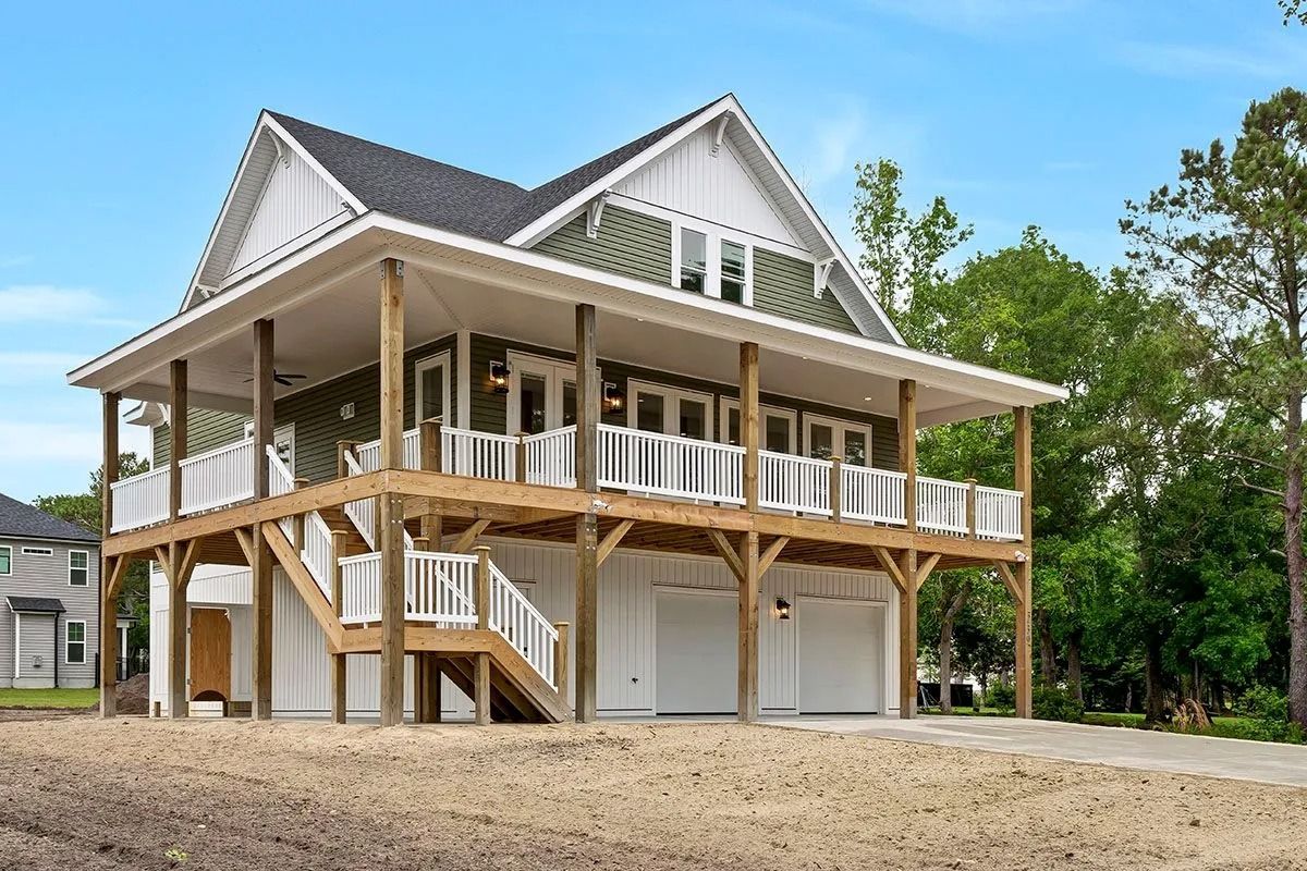 Two-story green and white house with large porch and garage on a sandy lot.