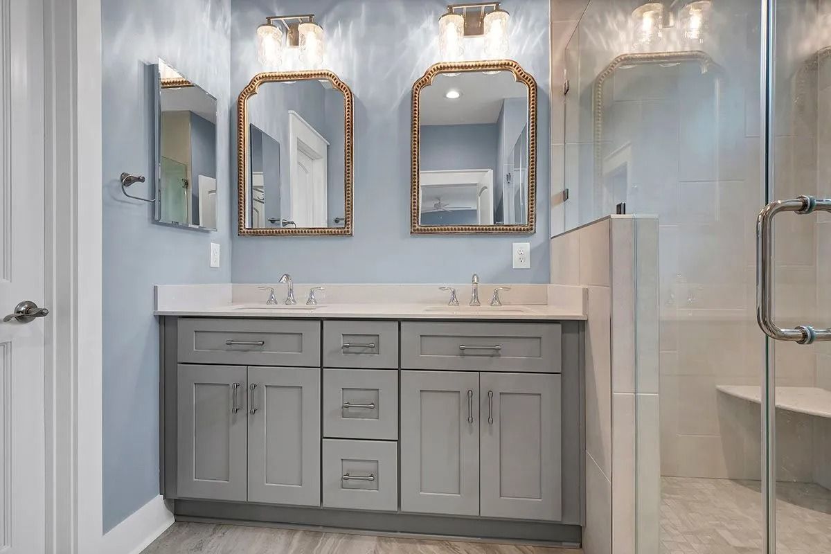 Light blue bathroom with double sinks, gray cabinets, gold mirrors, and a glass shower.