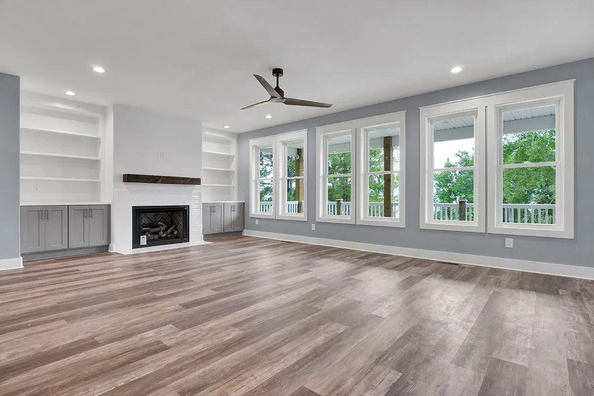 Empty living room with fireplace, built-in shelves, and large windows; light blue walls, wood floor.