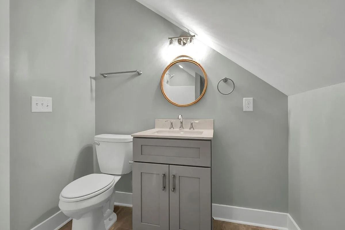 Bathroom with gray walls, toilet, vanity, and oval mirror under sloped ceiling.