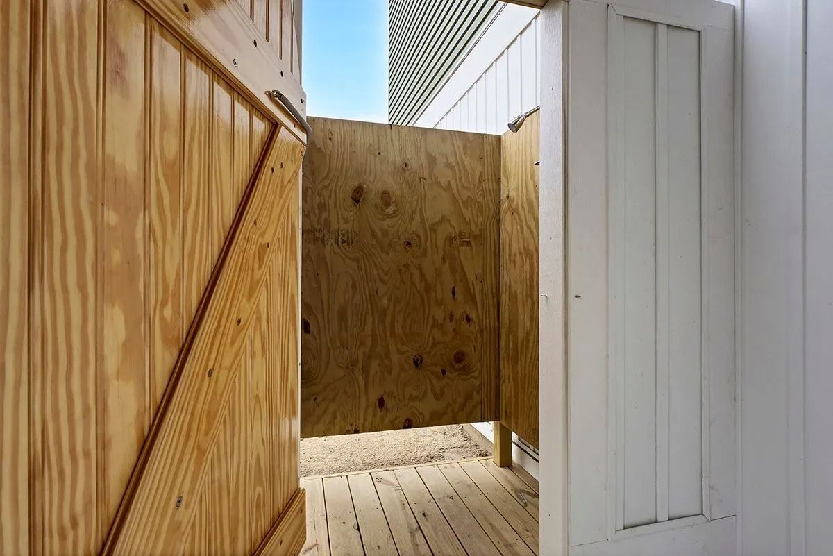 Open-air beach shower with wooden walls and door, sunlit interior.