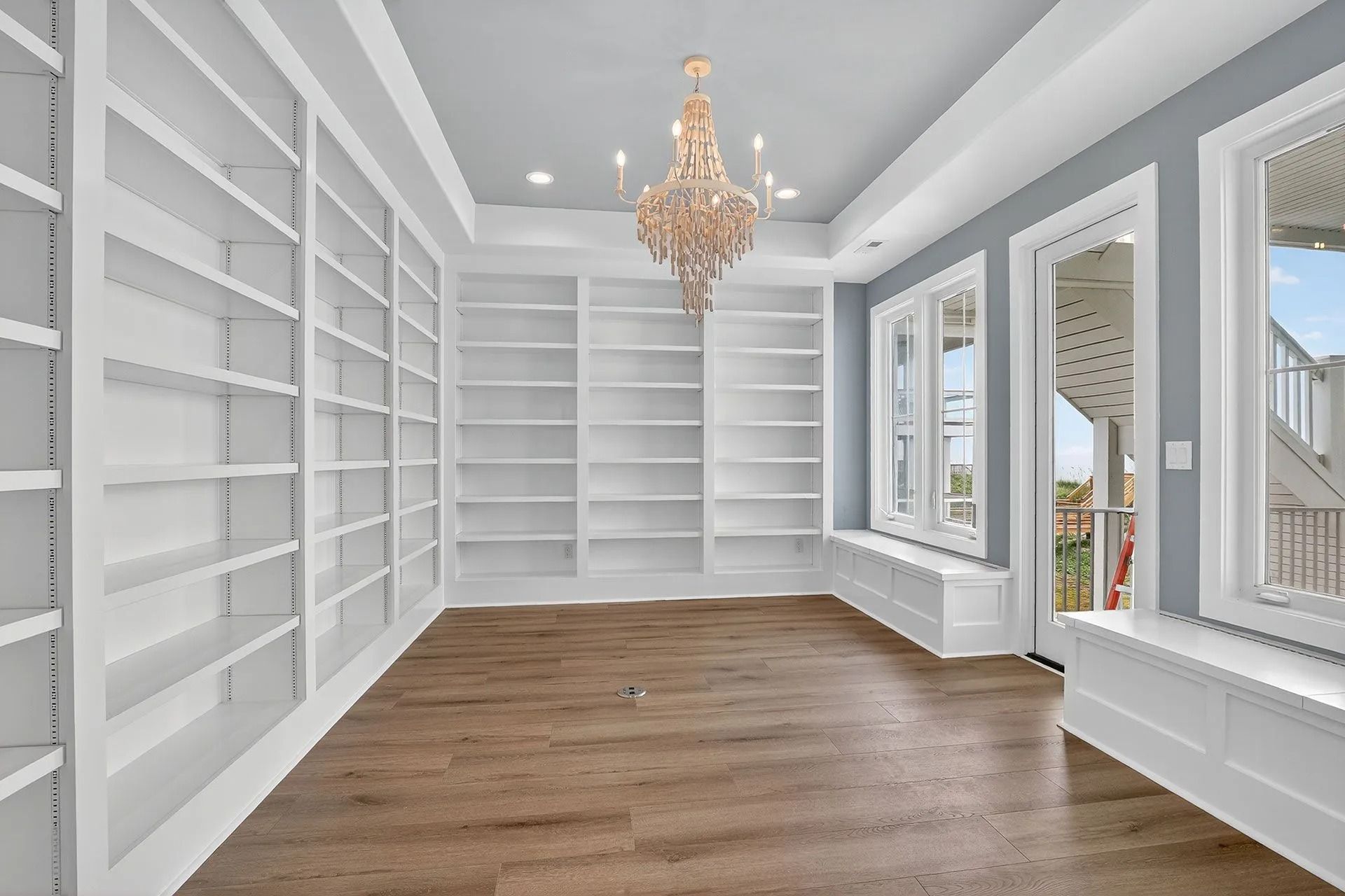 Empty white library with built-in shelves, wood floor, and chandelier, with windows overlooking a deck.