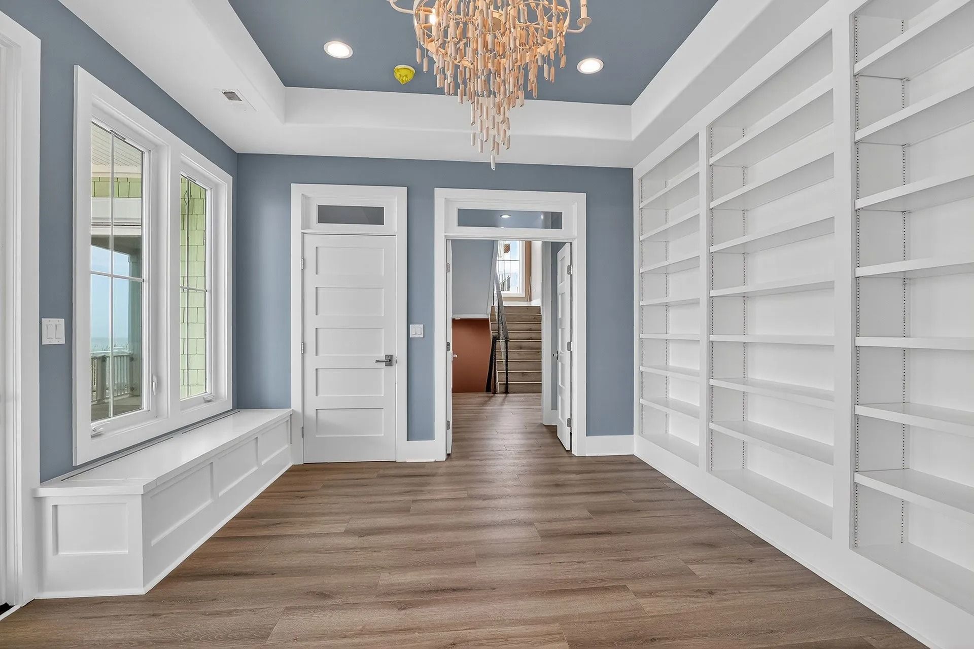 Blue-walled room with built-in bookshelves, window seat, doorway, and chandelier; wood flooring.