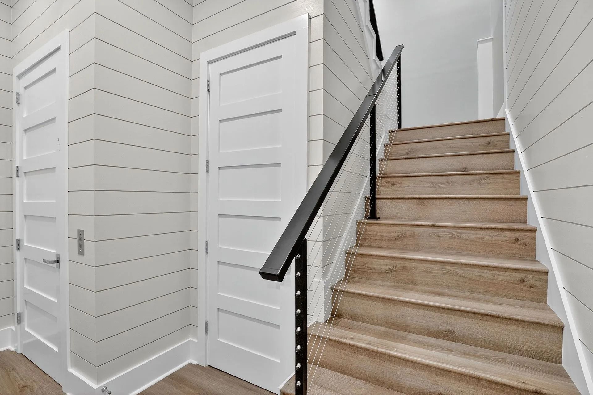 Interior view: white doors, wooden staircase with black railing, white shiplap walls.