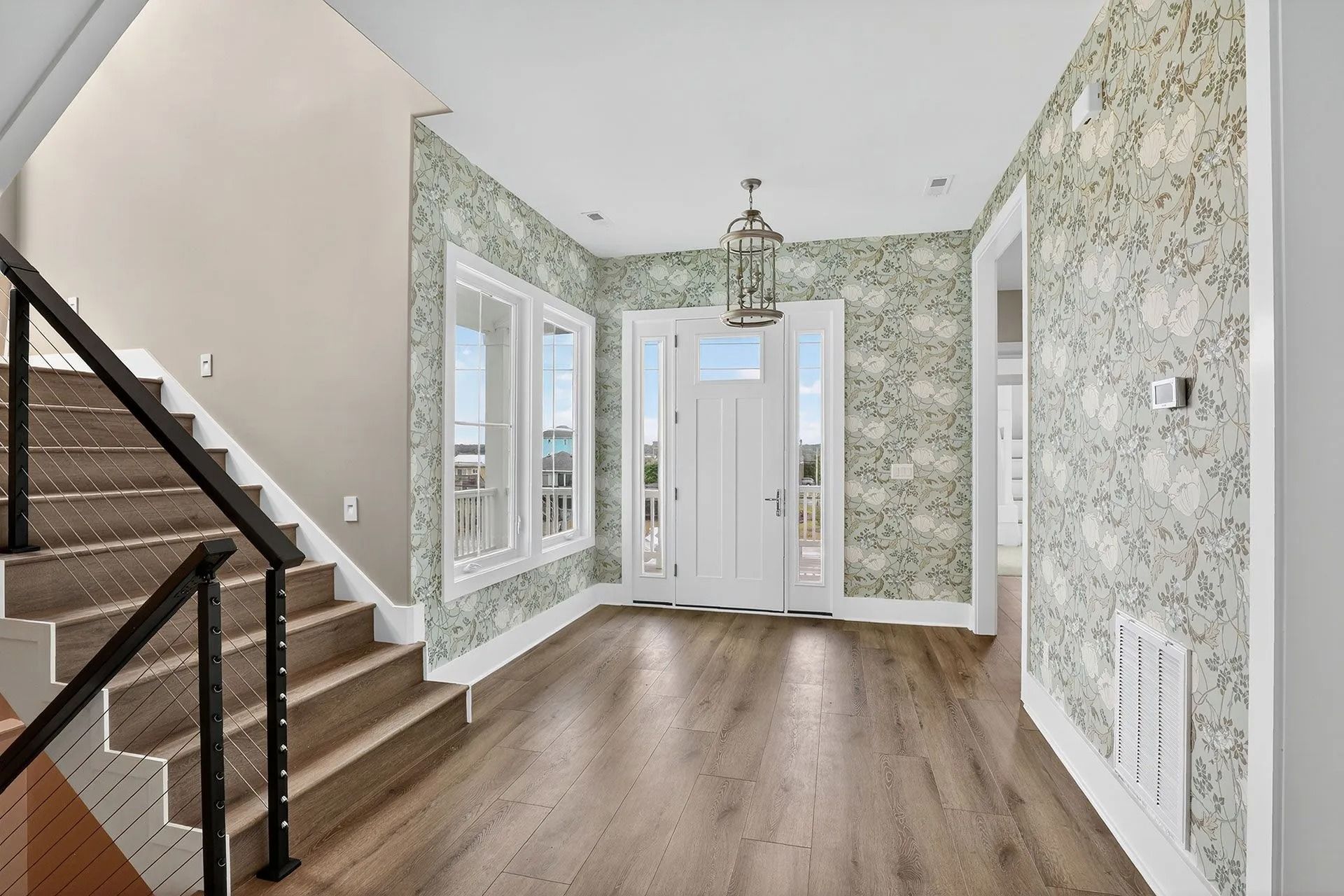 Entryway with staircase, floral wallpaper, and wood flooring. A door with sidelights is at the end of the hall.