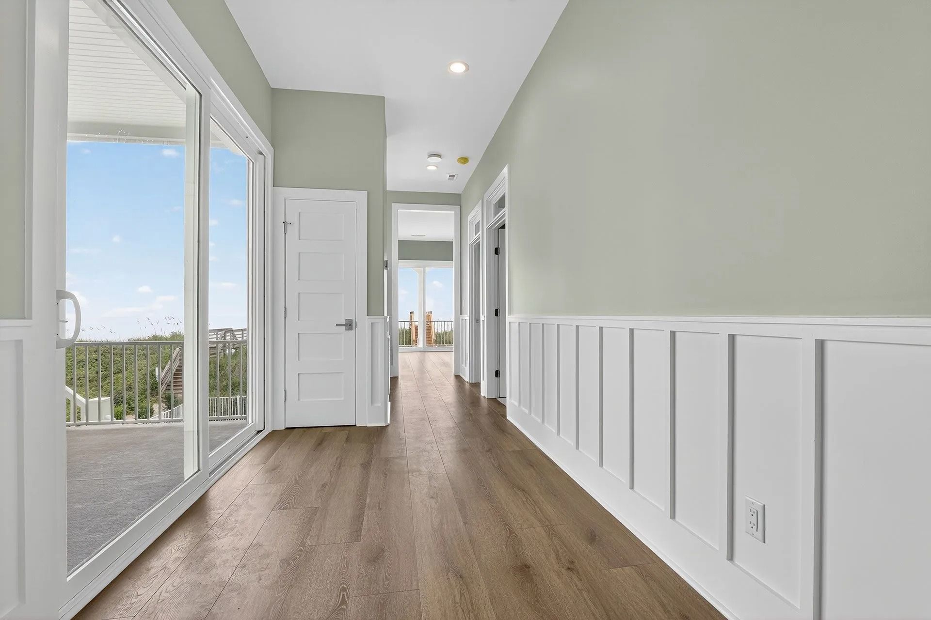 Long hallway with wood floors, light green walls, white wainscoting and trim, and large sliding glass door.