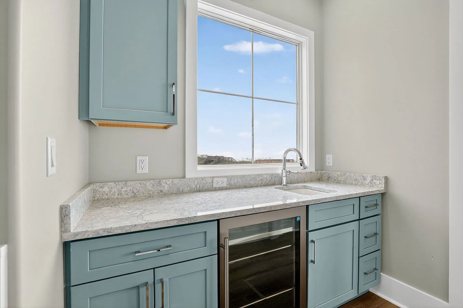 A small kitchen area with blue cabinets, a countertop, sink, and wine fridge under a window.