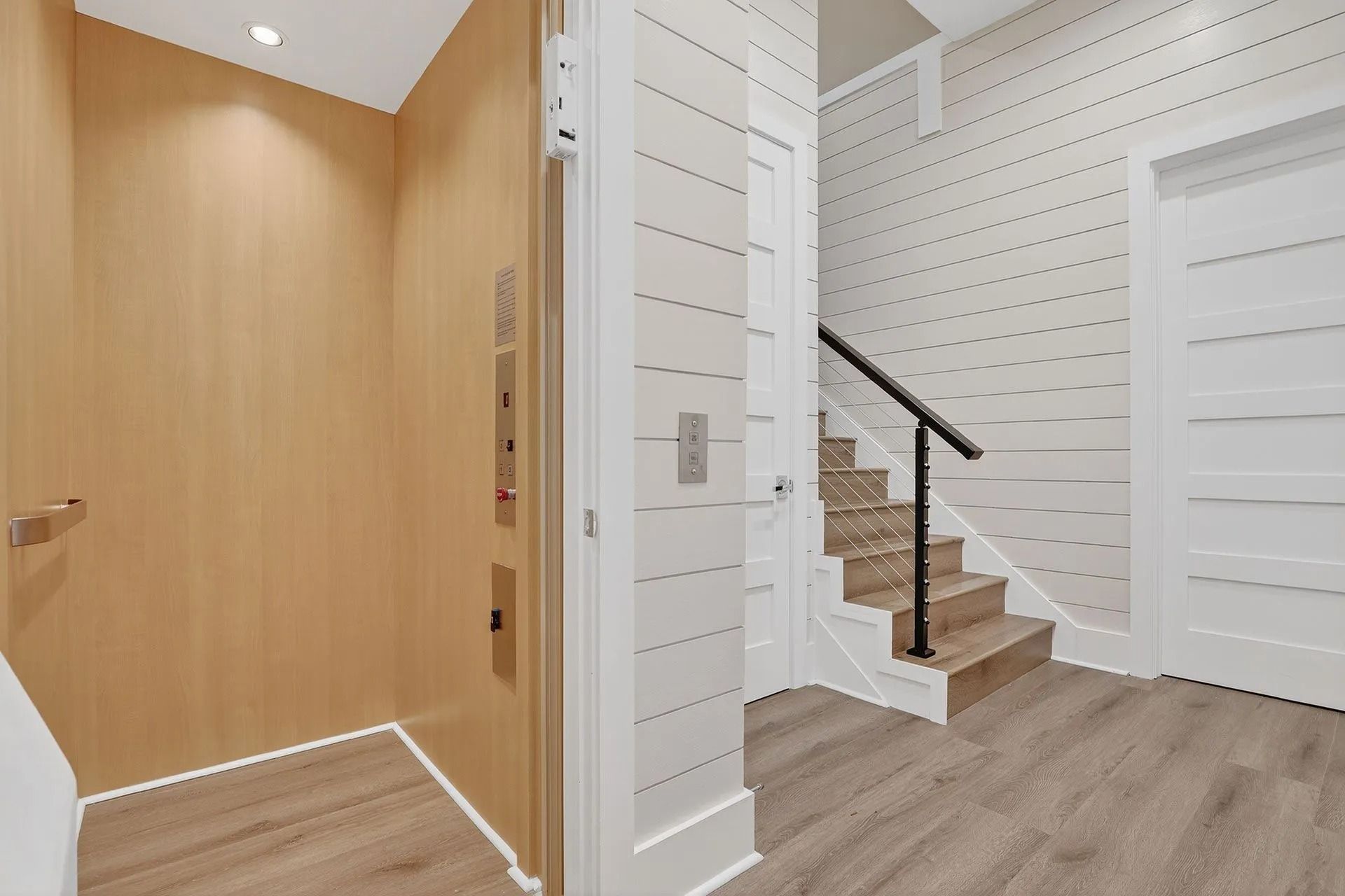 Hallway with elevator and stairs, light wood paneling, white walls, and wood-look flooring.