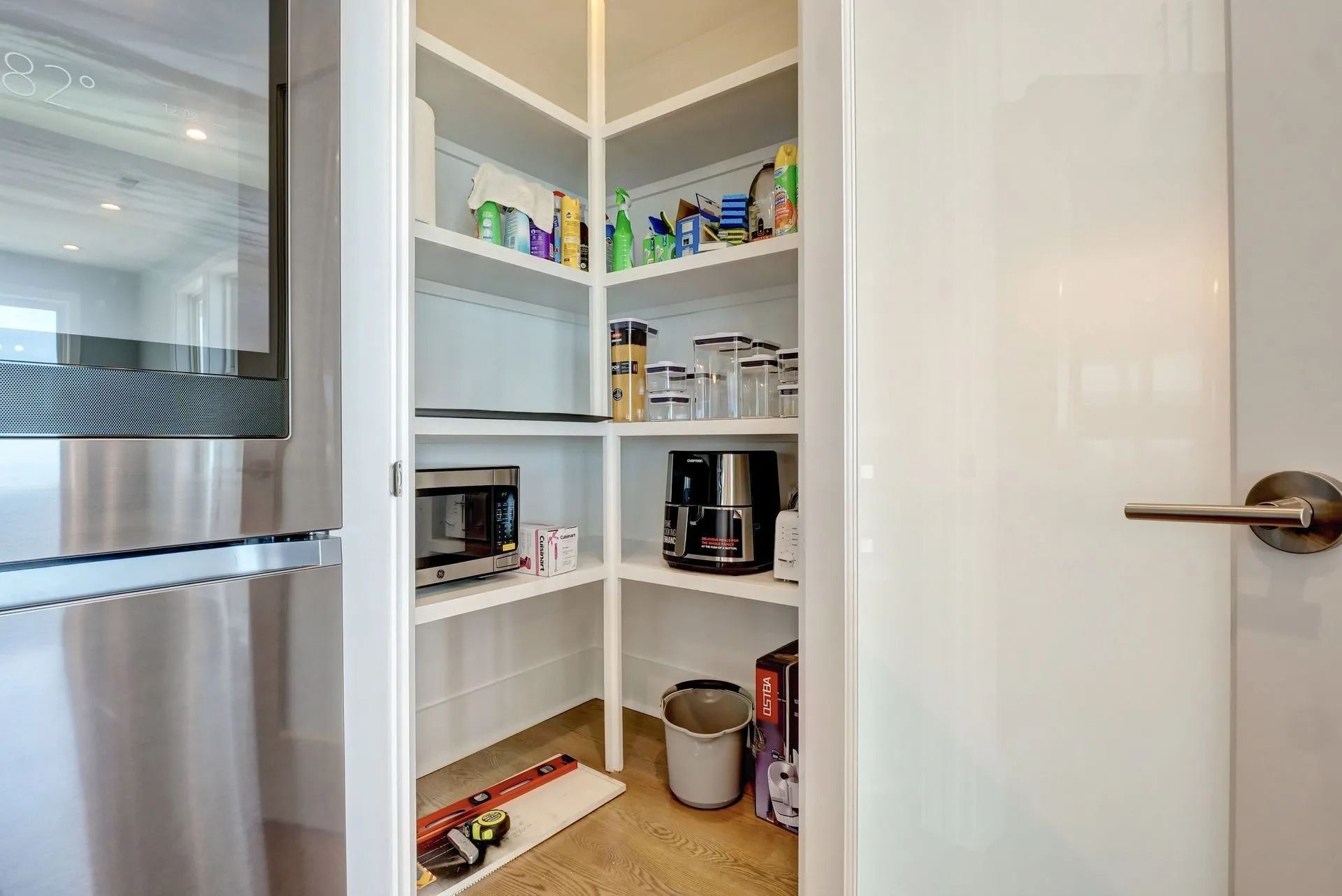 White pantry with shelves holding food, cleaning supplies, and small appliances next to a refrigerator.