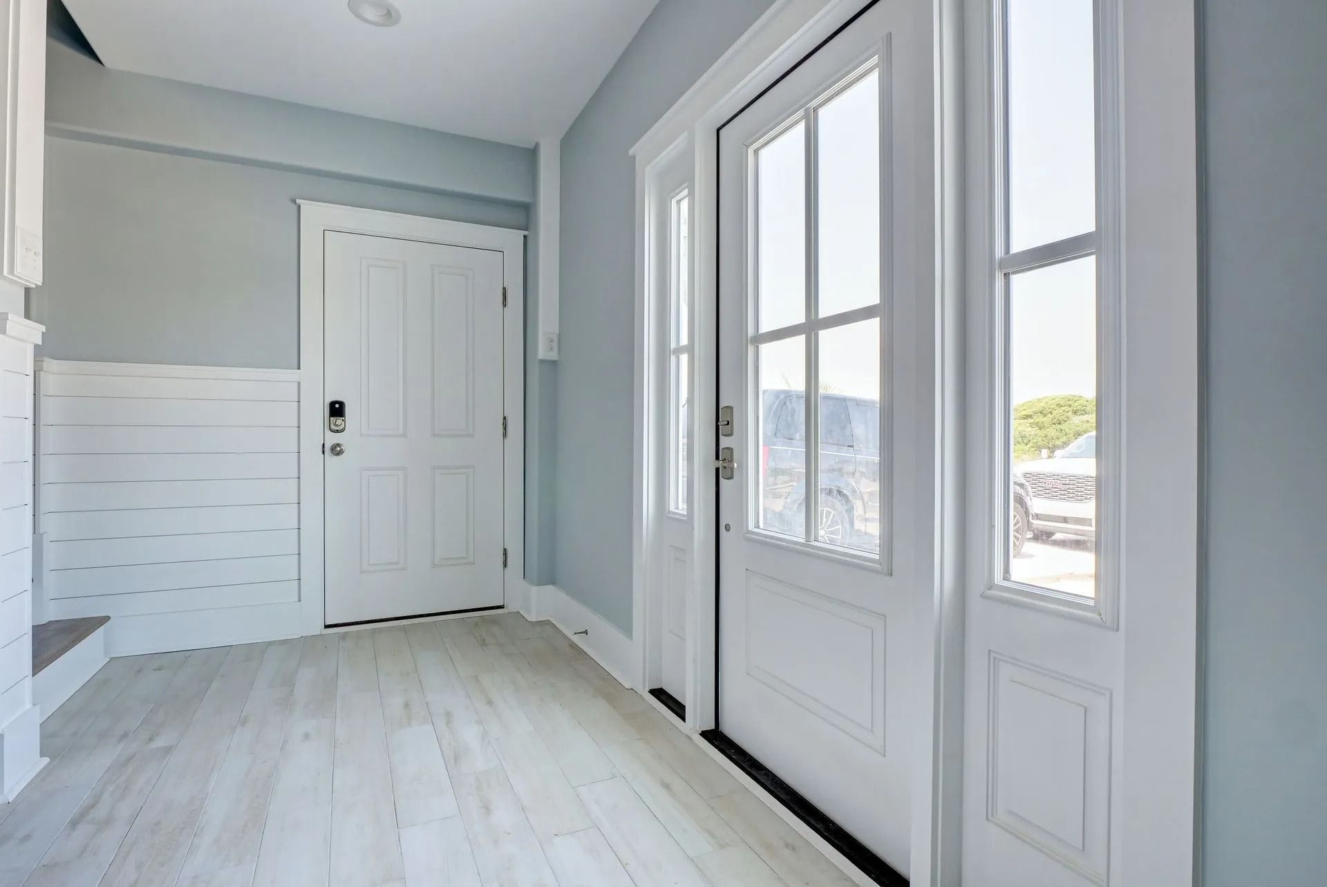Bright entryway with white doors and trim, light wood floors, and blue-gray walls. Sunlight streams in.