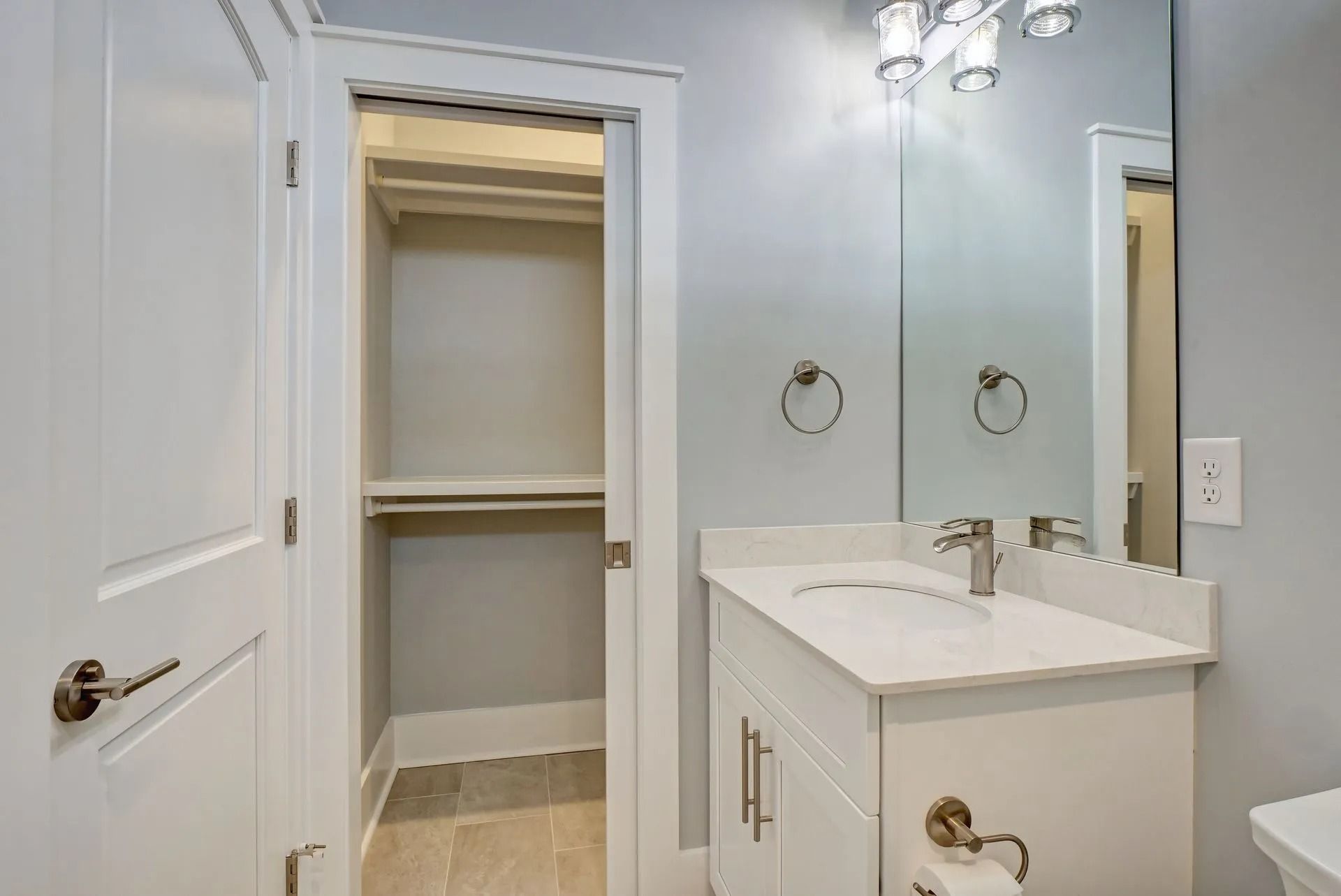 White bathroom with a closet, vanity, mirror, and a toilet paper dispenser.