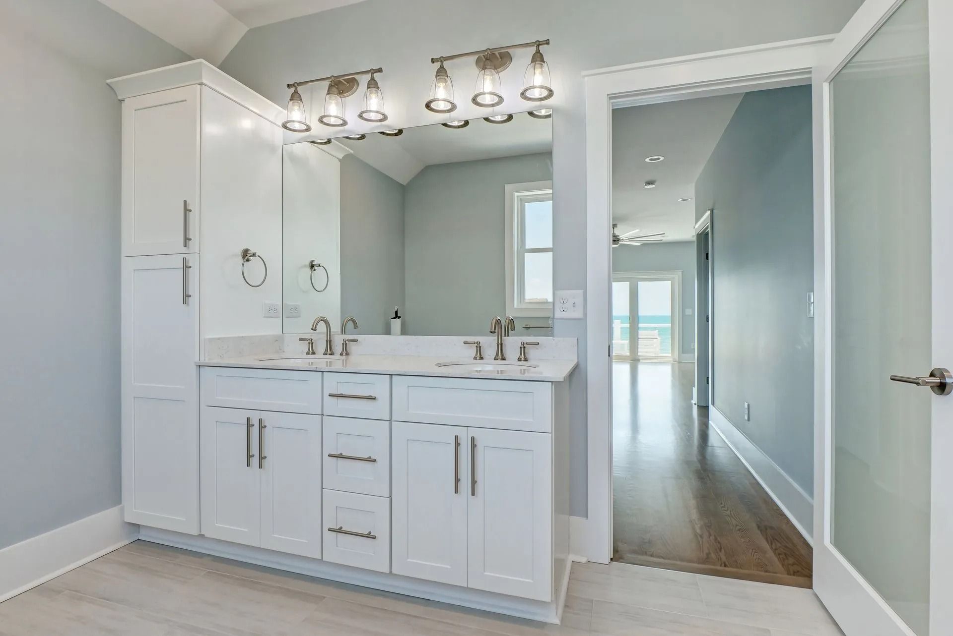 White bathroom vanity with double sinks, mirror, and tall storage cabinets. Doorway leads to hallway with ocean view.
