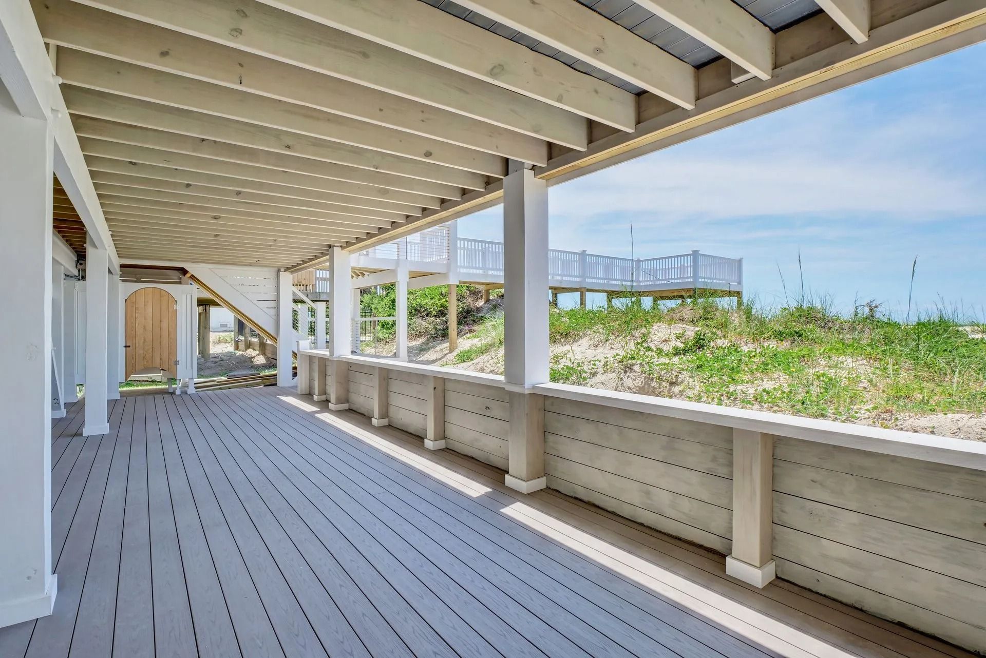 Covered wooden deck overlooking sand dunes and the ocean, with a distant deck.
