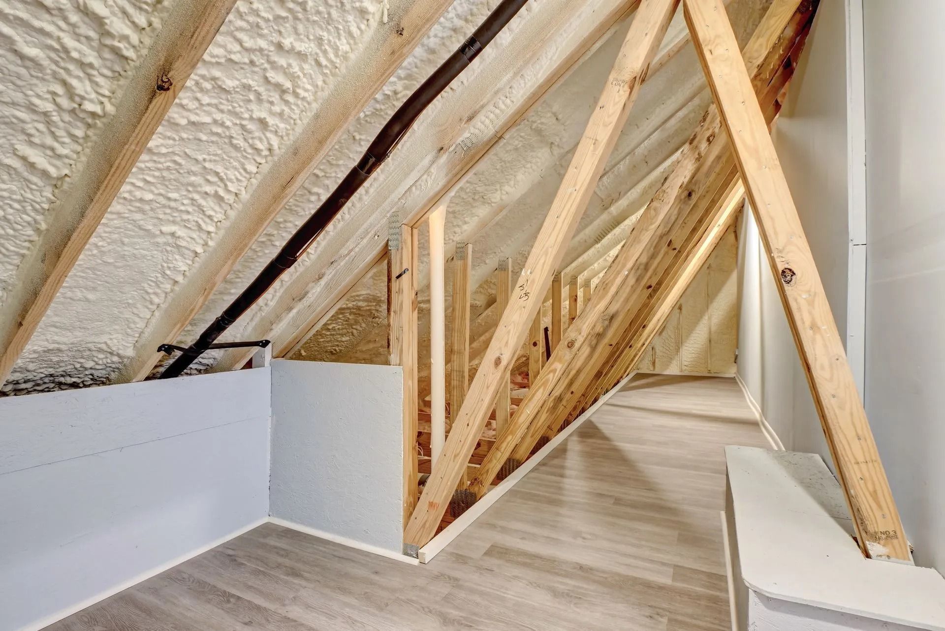 Interior view of an attic space with wooden rafters, spray foam insulation, and gray walls and flooring.