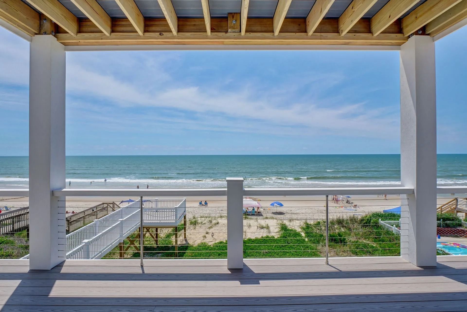 Ocean view from a porch with white columns, overlooking a beach with people, under a blue sky.