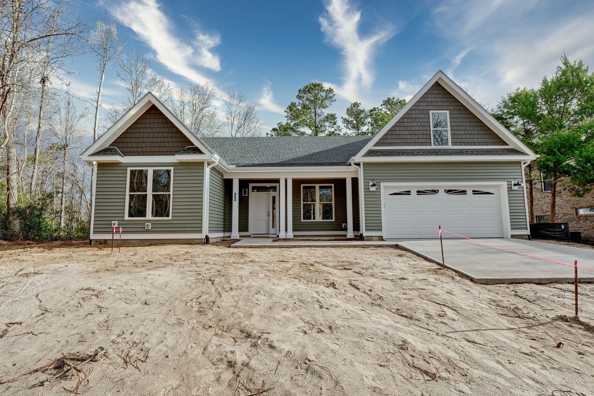 Green house with white garage door and concrete driveway on sandy lot under blue sky.