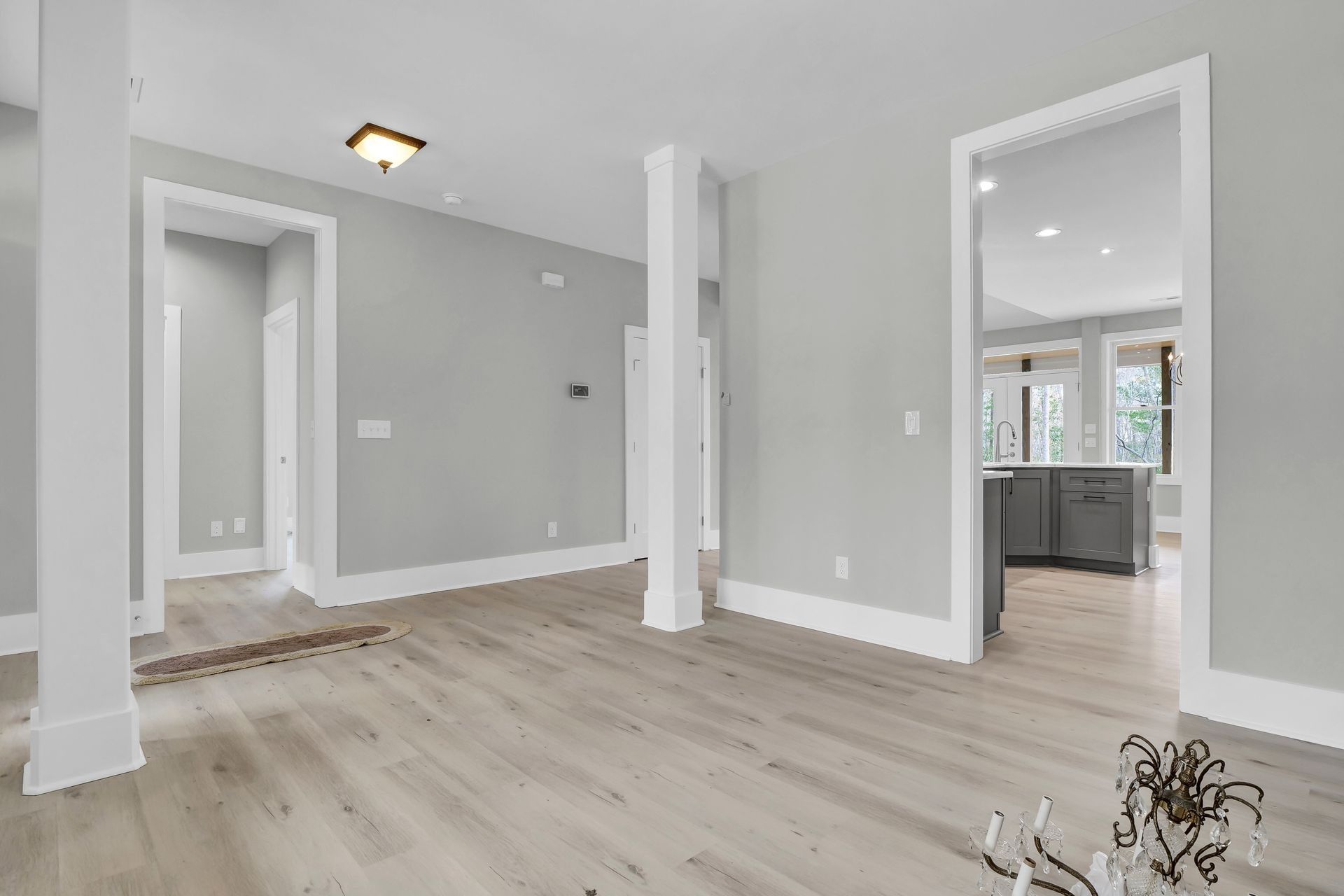 Empty room with light gray walls, white trim, and light wood flooring. Kitchen visible in the background.