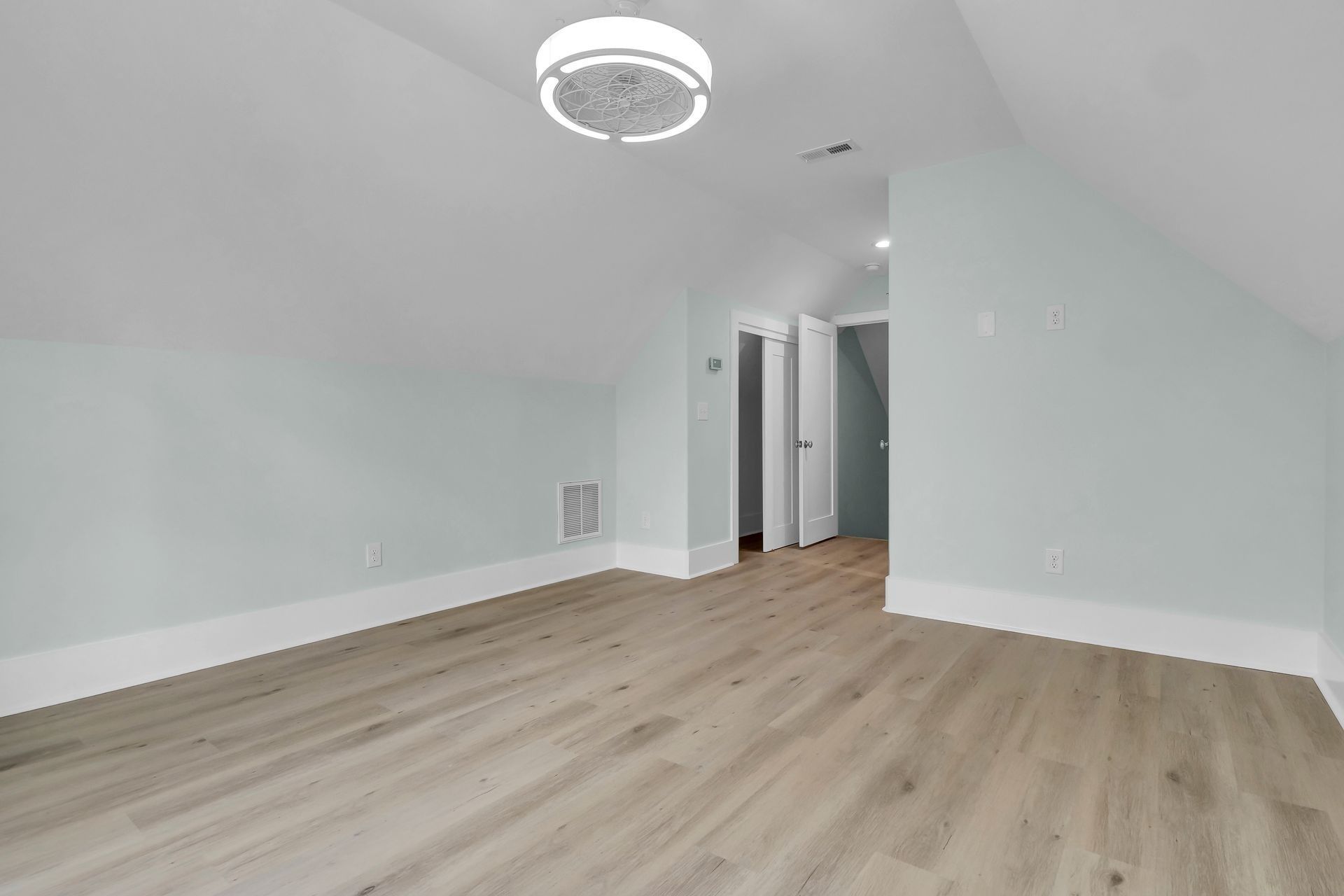Empty room with slanted walls, light blue paint, light wood floors, and a modern ceiling light.