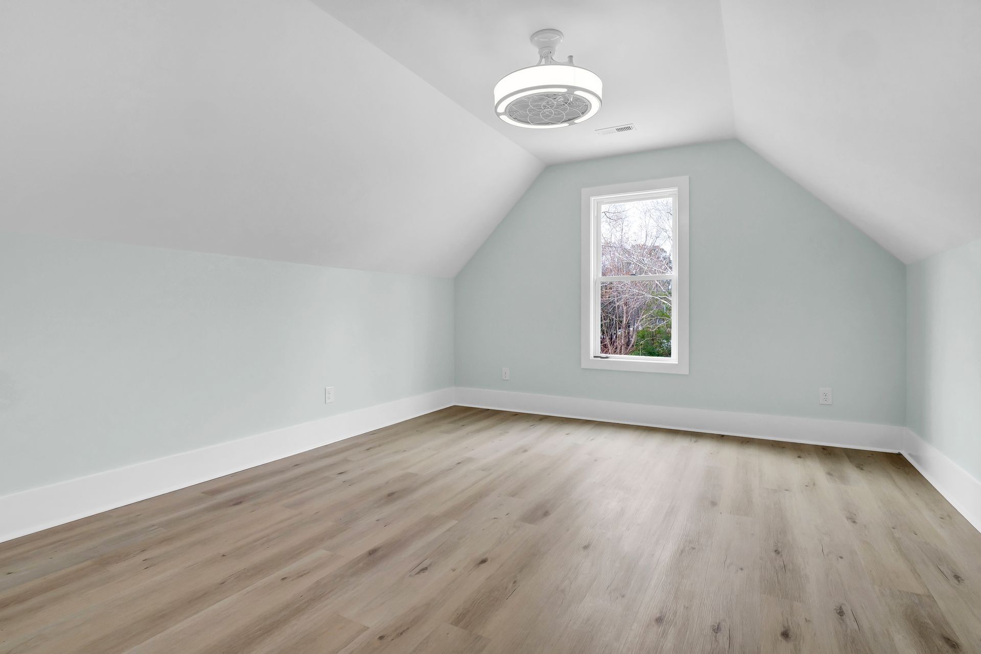 Empty attic room with sloped ceiling, window, wood-look floor, and pale blue walls.