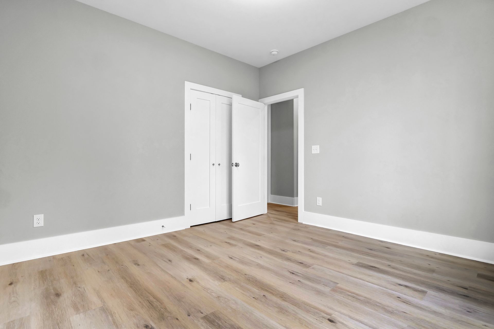 Empty room with gray walls, white trim, and wood-look flooring. Closet with closed doors, doorway.