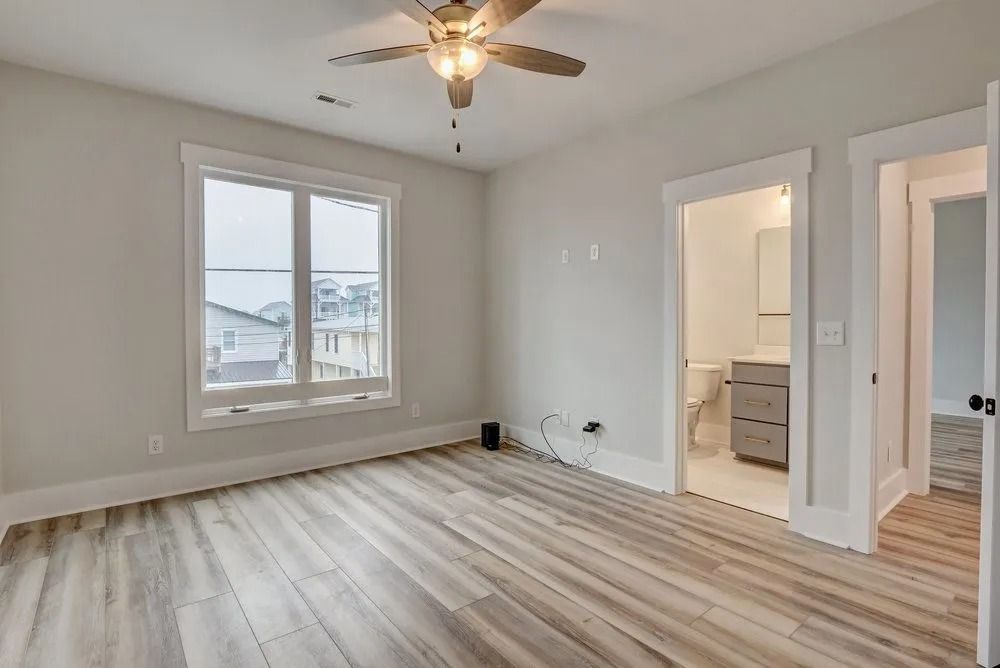 Empty bedroom with light wood floors, a large window, and an open doorway to a bathroom.