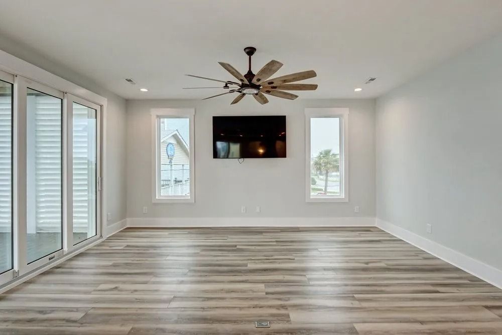 Empty living room with wood-look flooring, light blue walls, a ceiling fan, TV, and large windows.