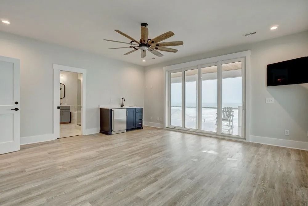 Empty room with bar area, large windows, ceiling fan, TV, and light wood flooring.