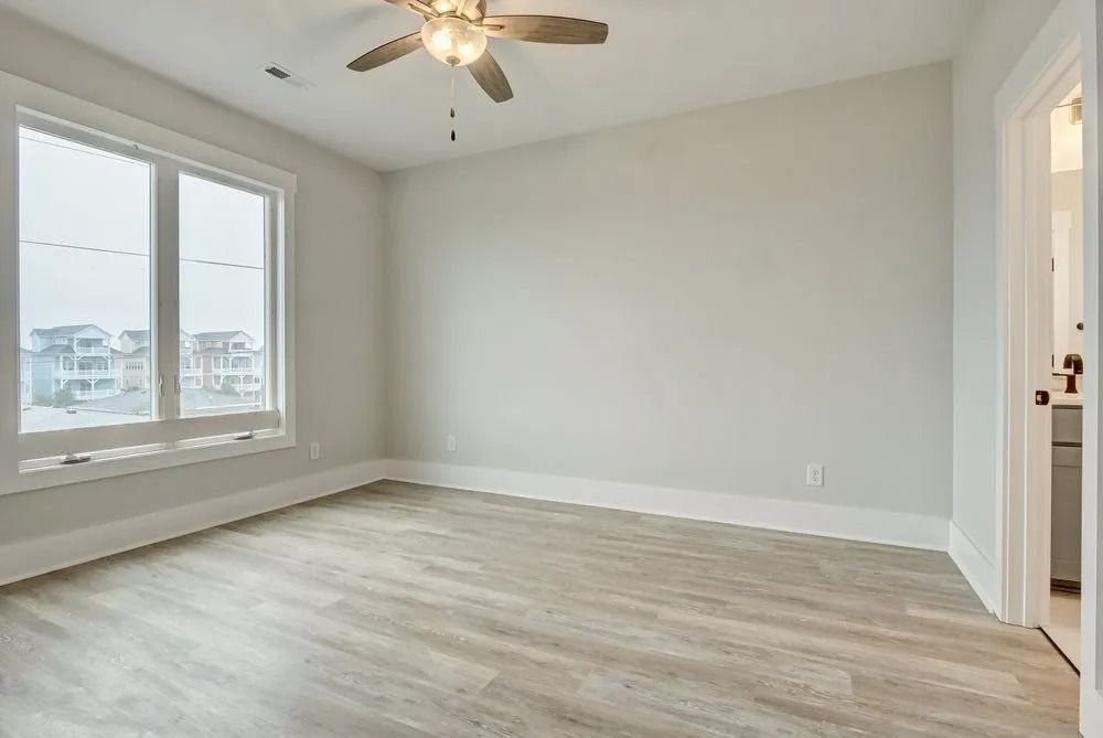 Empty bedroom with light wood flooring, large window, and light gray walls.