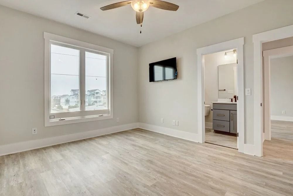 Empty bedroom with large window, TV, and open doorway to a bathroom. Light gray walls and flooring.