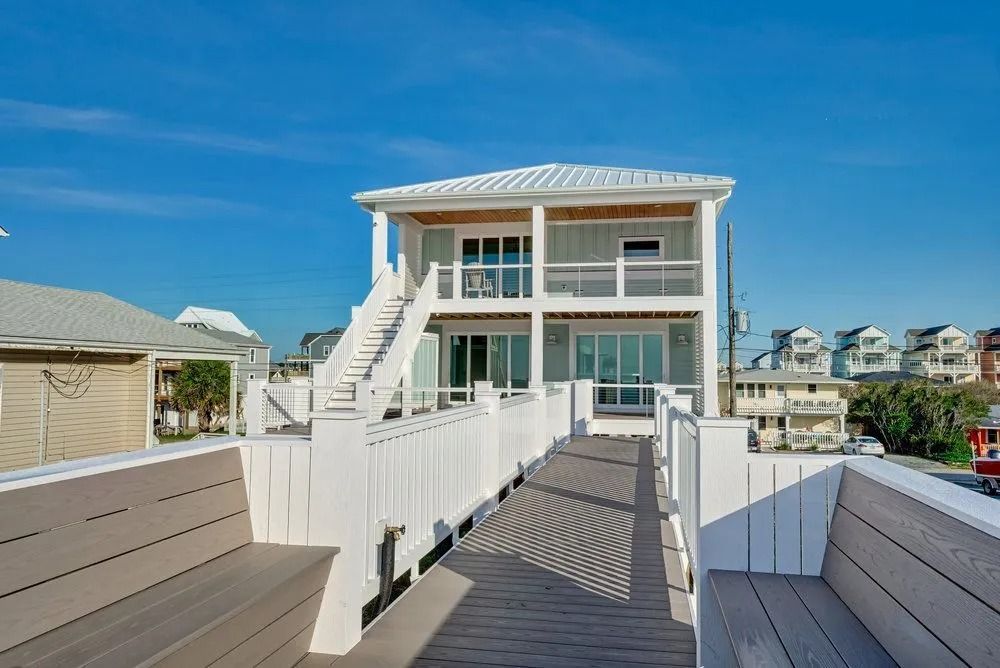 Two-story beach house with a white exterior, balcony, and staircase leading up to it, clear blue sky.